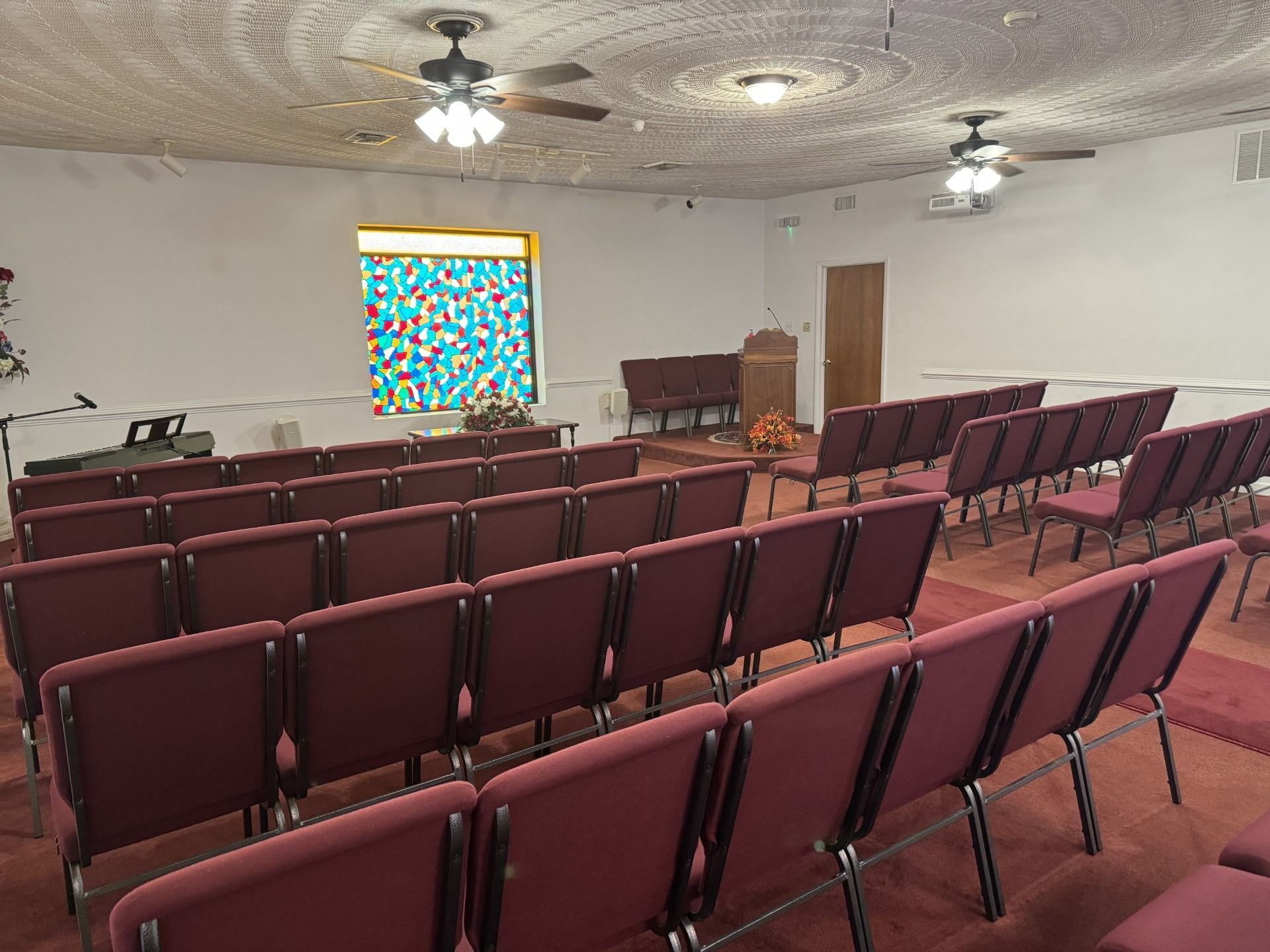 Rows of red chairs face a podium and stained glass window in a church.
