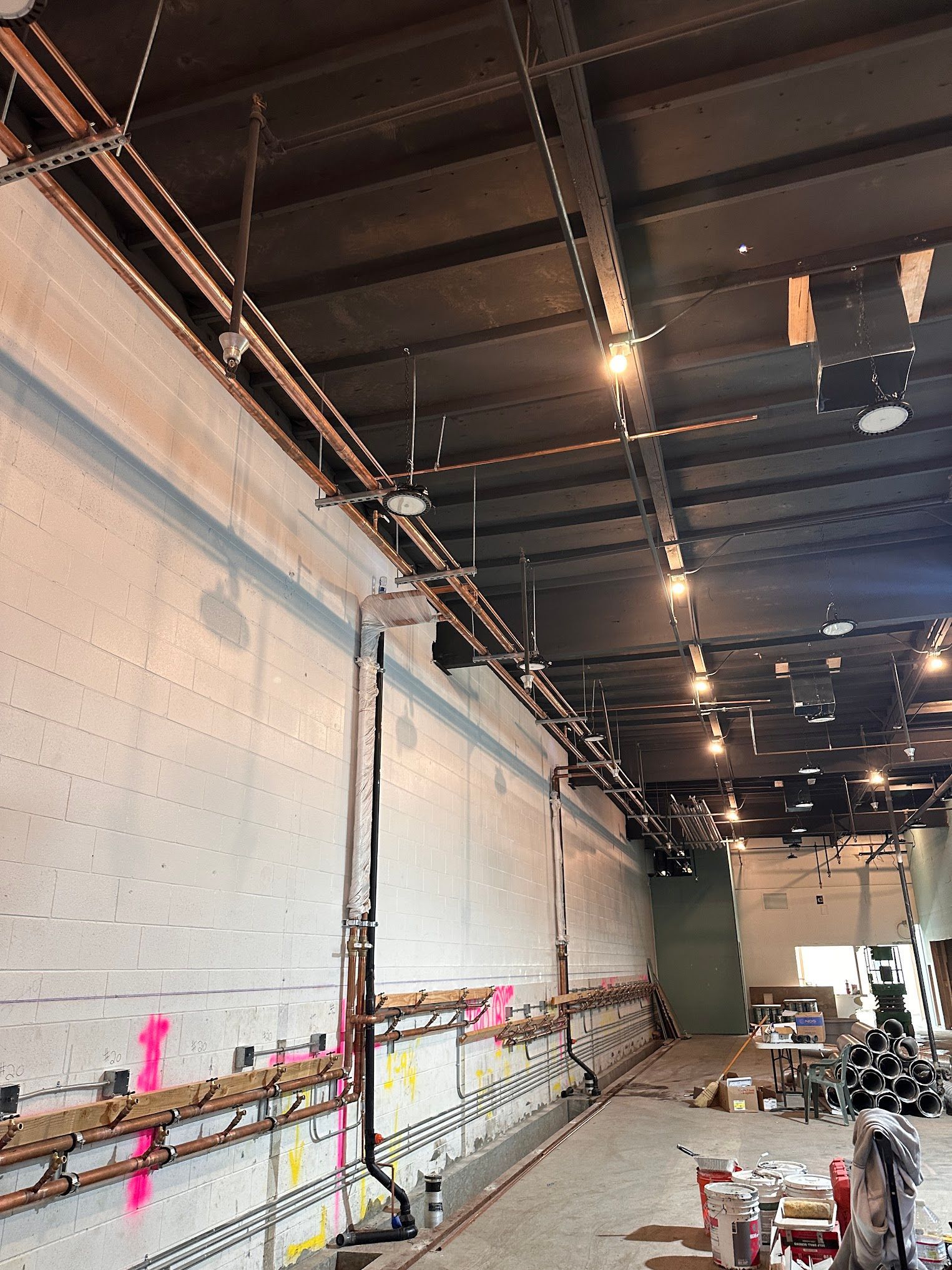 Interior of a building under construction. Exposed ceiling beams and wiring, unfinished walls, copper pipes, and lights.