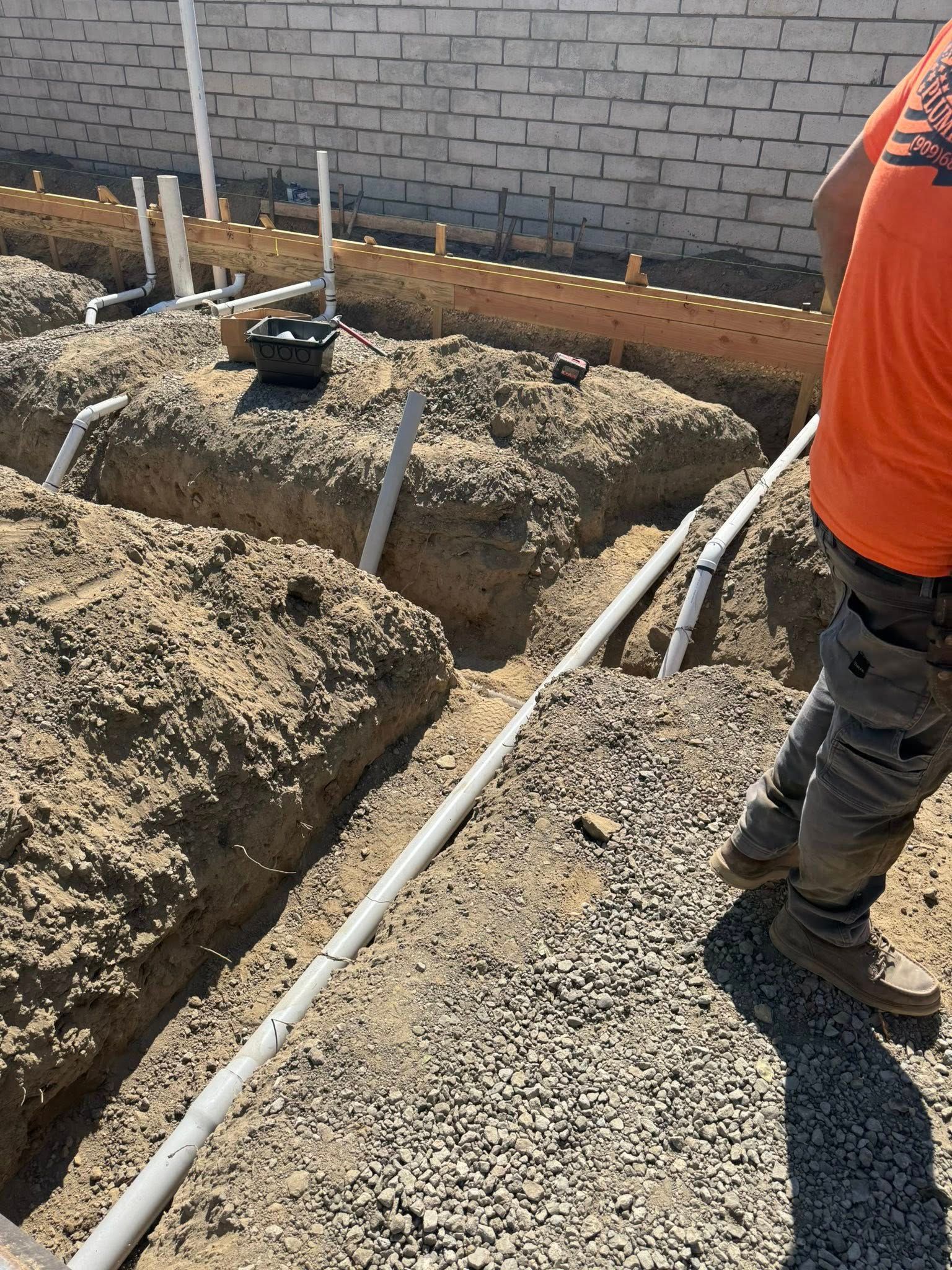 Plumber installing pipes in a trench at a construction site near a brick wall.