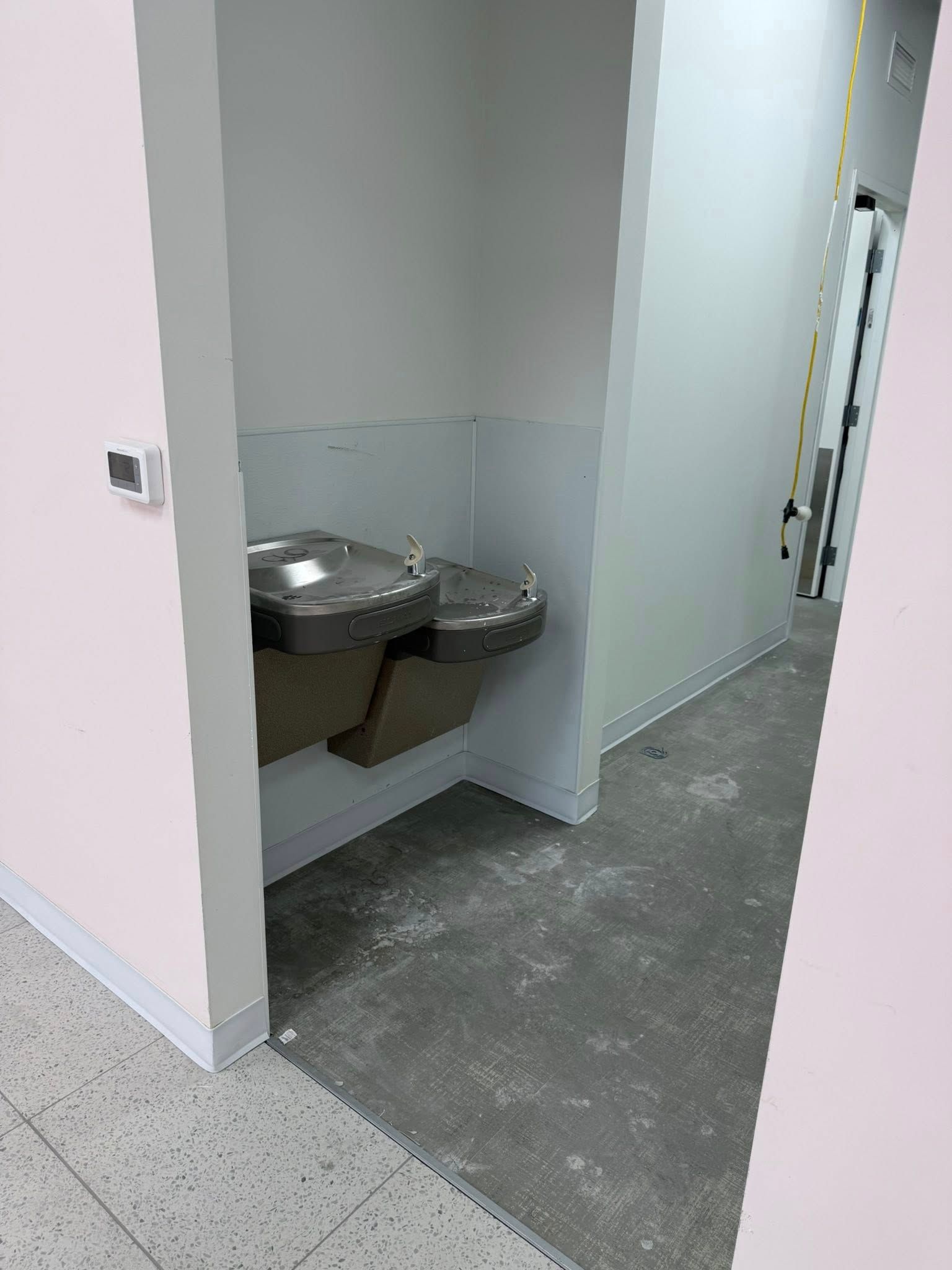 Water fountain in a hallway alcove. Stainless steel with two spouts. Gray concrete floor, white walls.