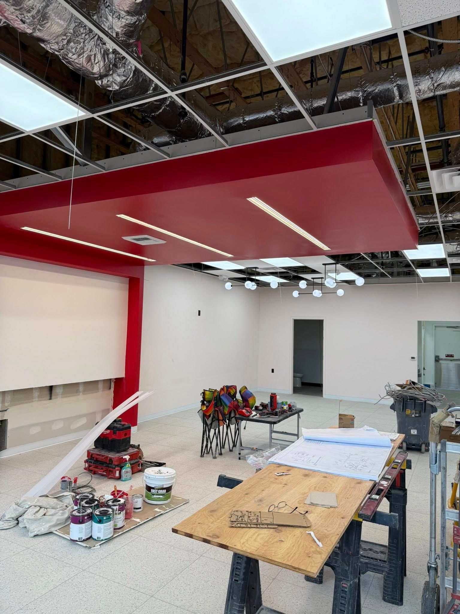 Interior construction site with red ceiling panel, tools, and plans. White walls, unfinished ceiling.