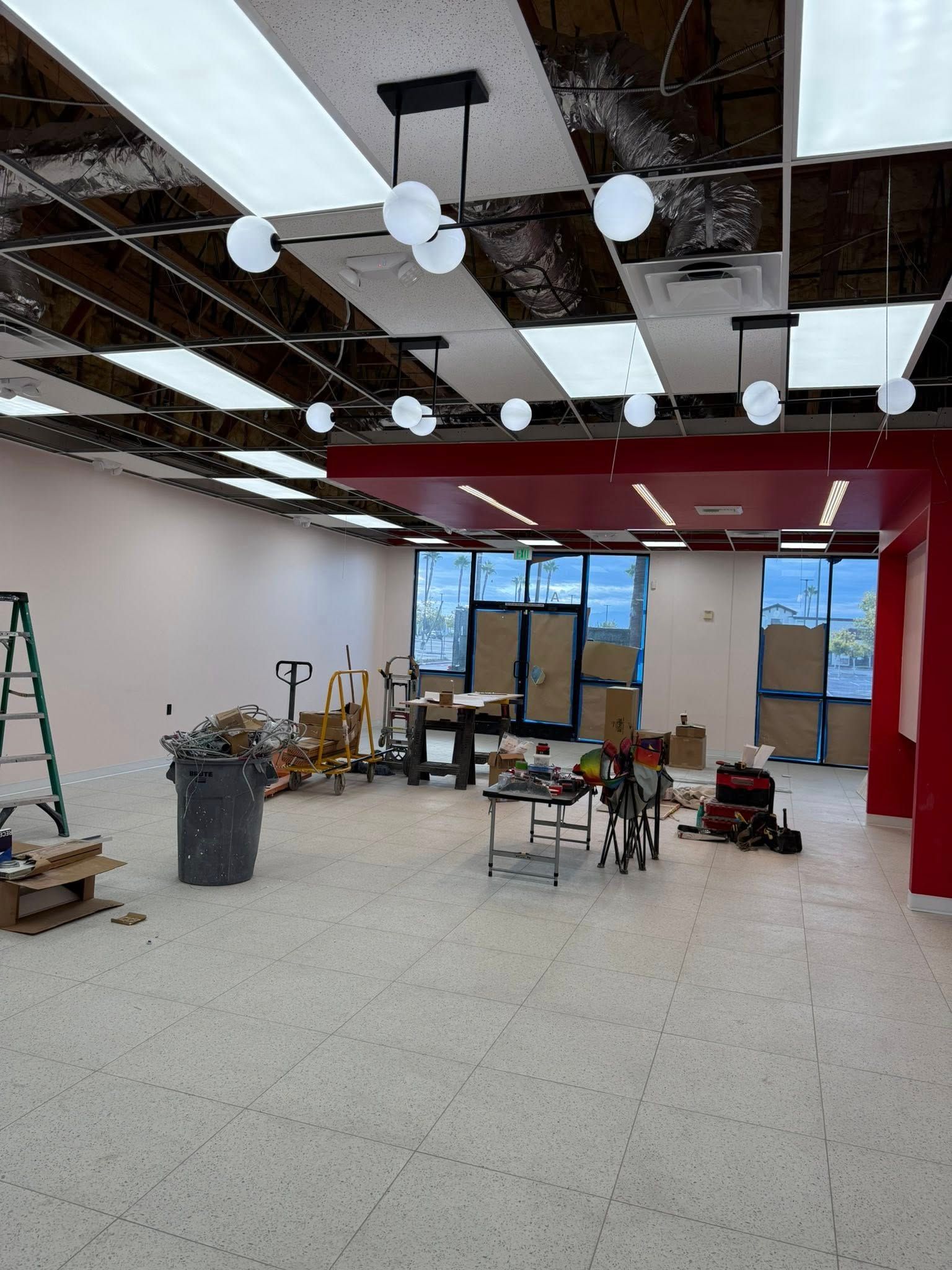 Interior construction site; open space with exposed ceiling, tools, debris, red accents, and large windows.