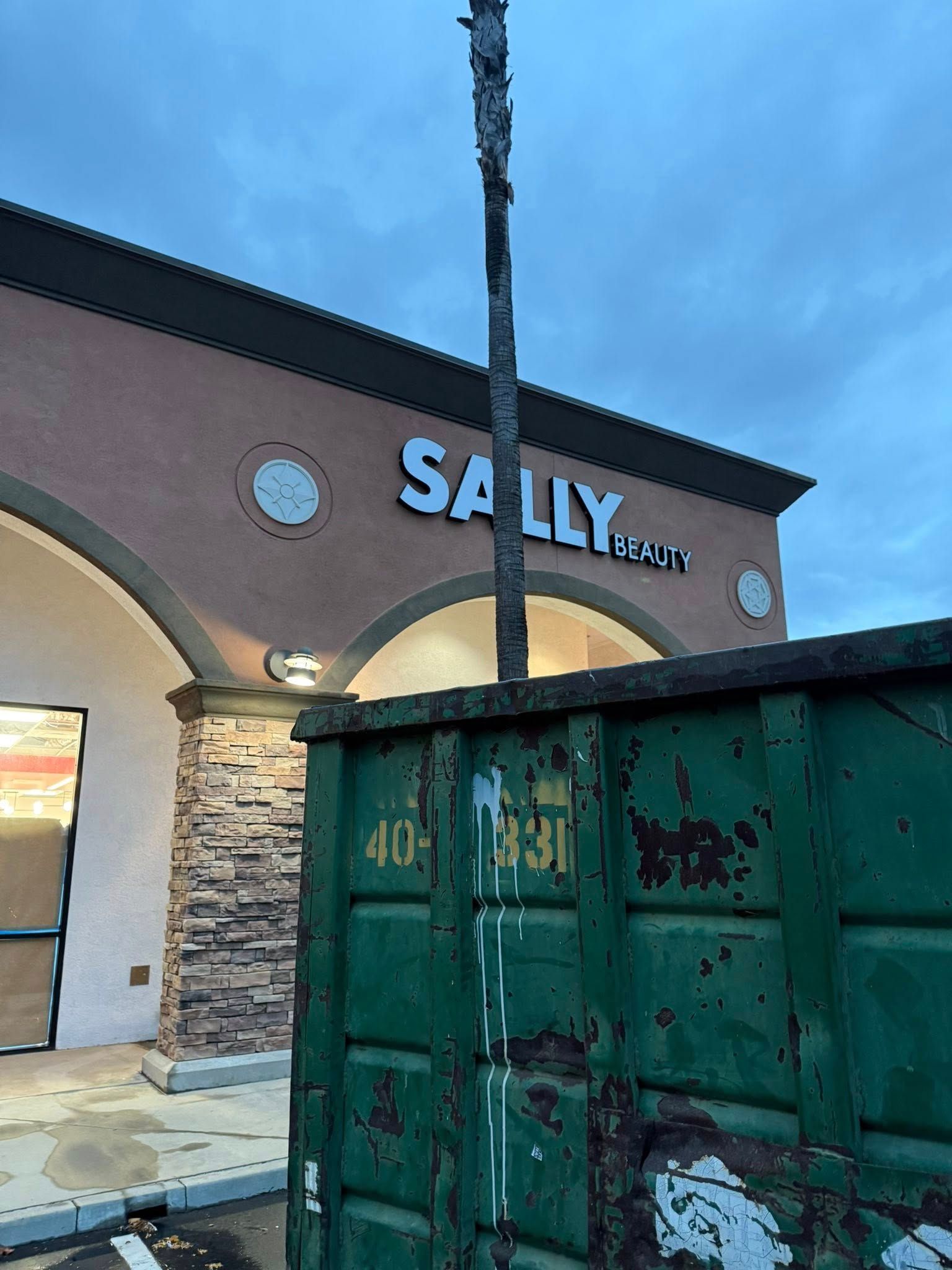 Green dumpster in front of a Sally Beauty store with arched entrance under a cloudy sky.