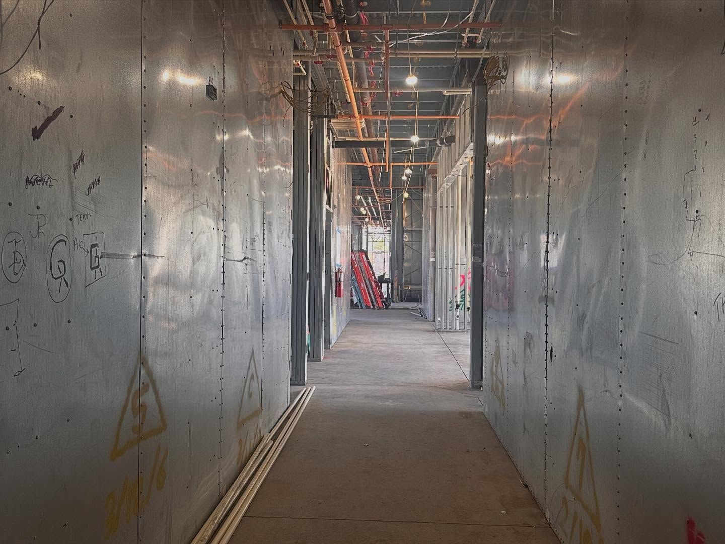 Narrow hallway with silver metal walls, under construction; pipes and wires overhead, tools at the end.