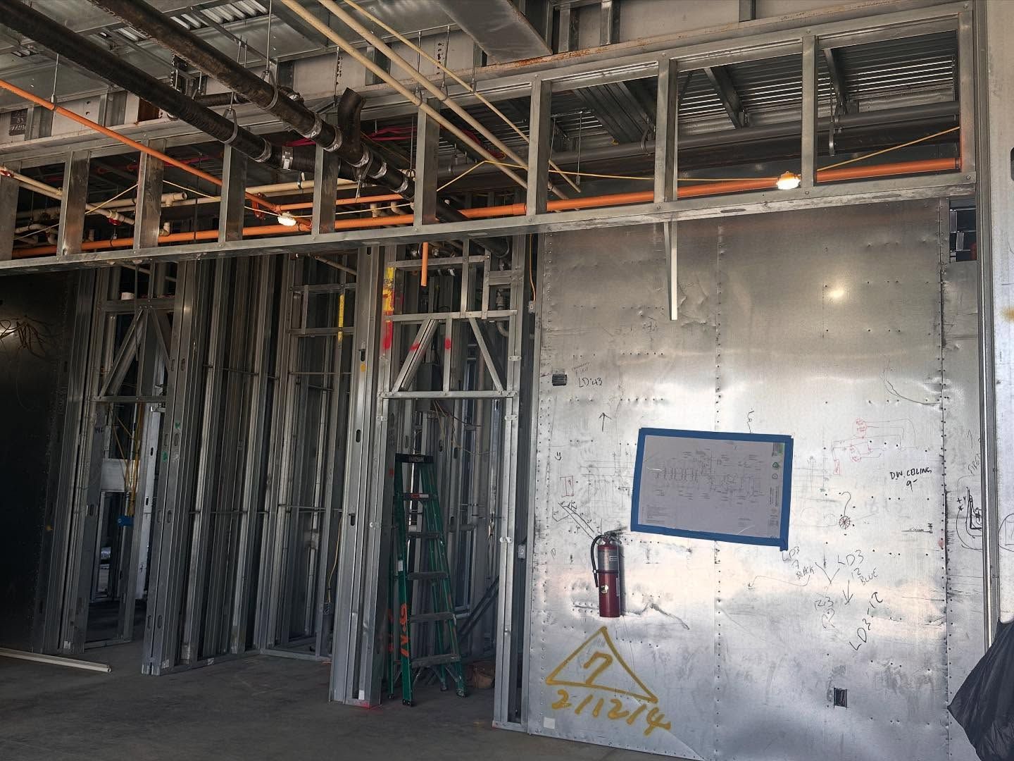 Construction site interior: metal framing, electrical conduits, and a sheet-metal wall, with a ladder.