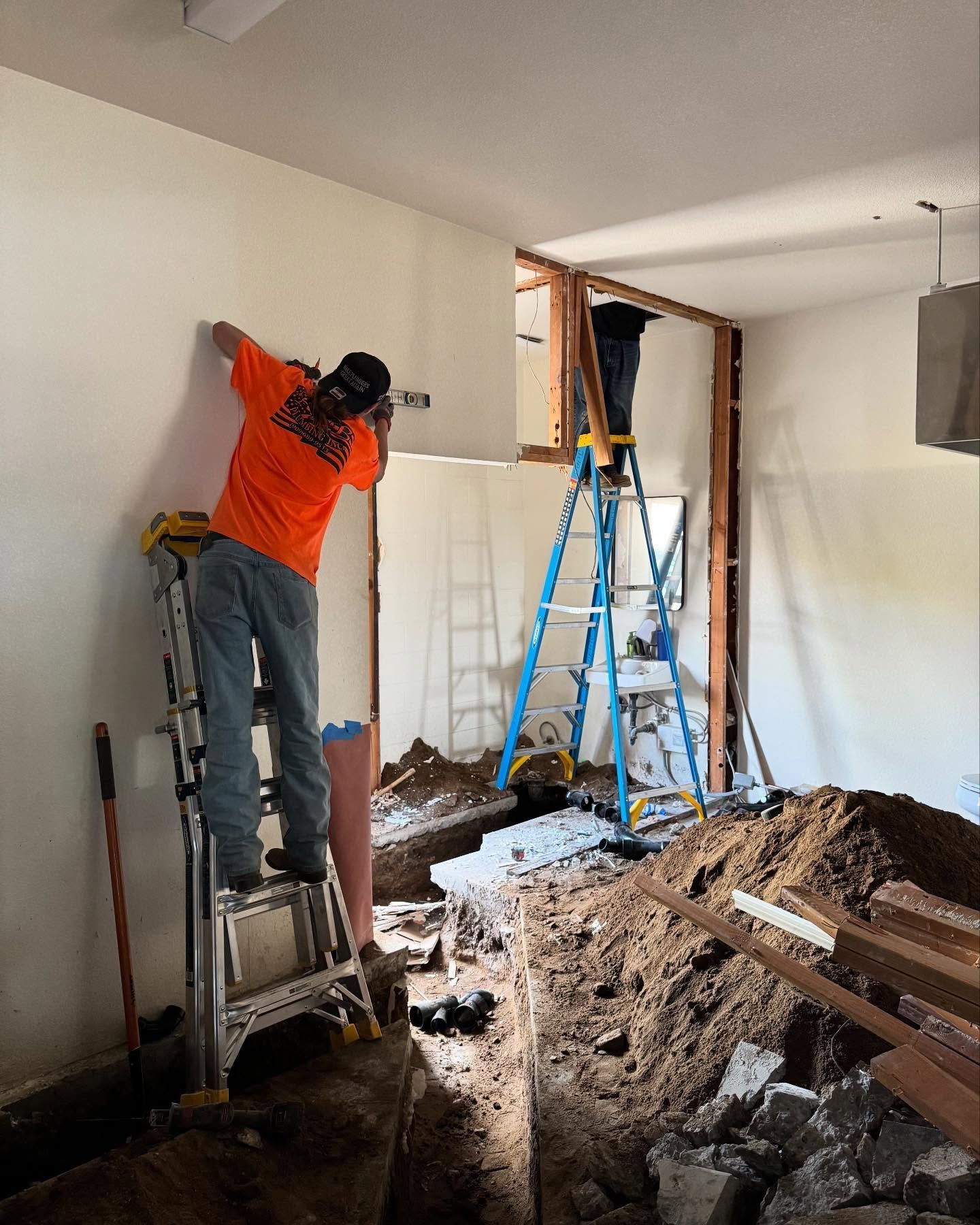 Construction worker on ladder, drilling into a wall. Interior demolition, with exposed framing and a trench.