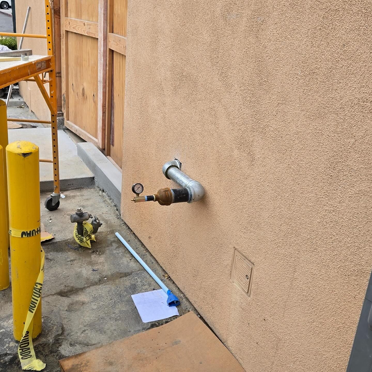 Tan stucco wall with a metal pipe and valve, yellow bollards, and construction materials.