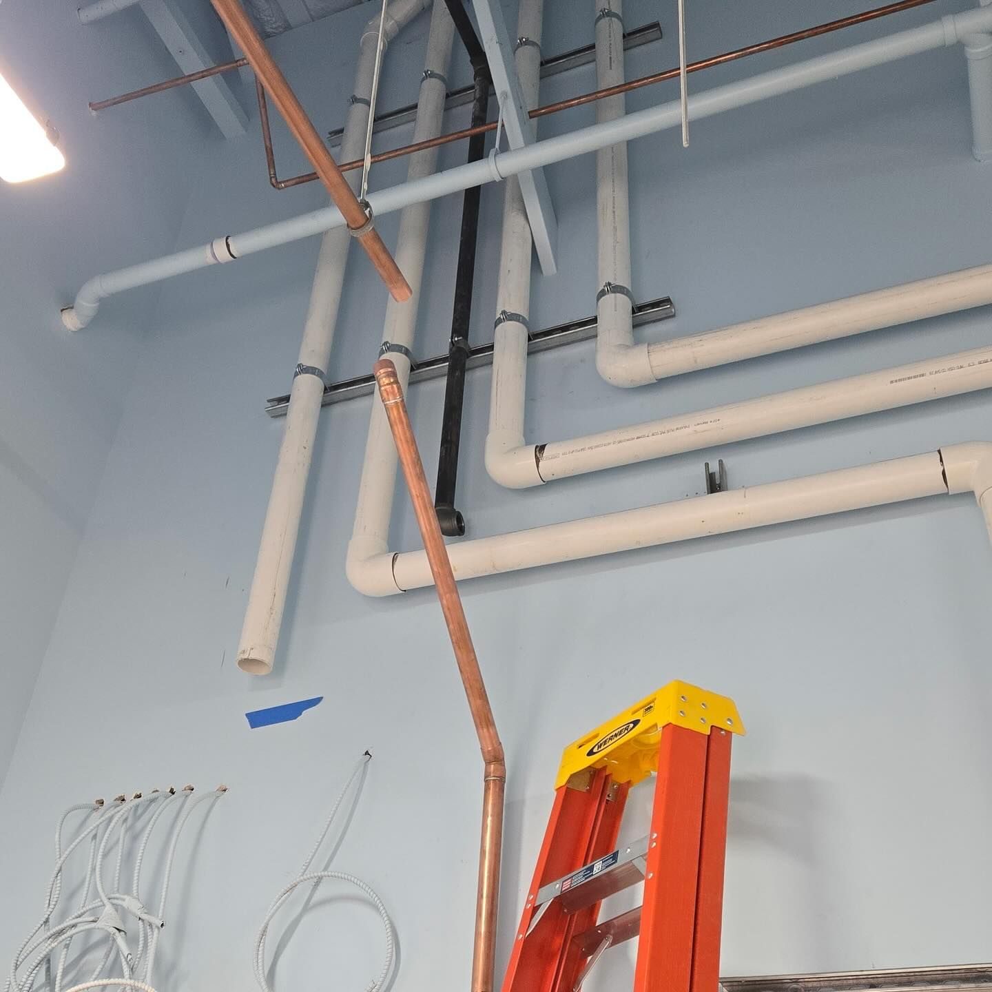 Plumbing pipes and a ladder against a light blue wall. Copper and white pipes are visible.