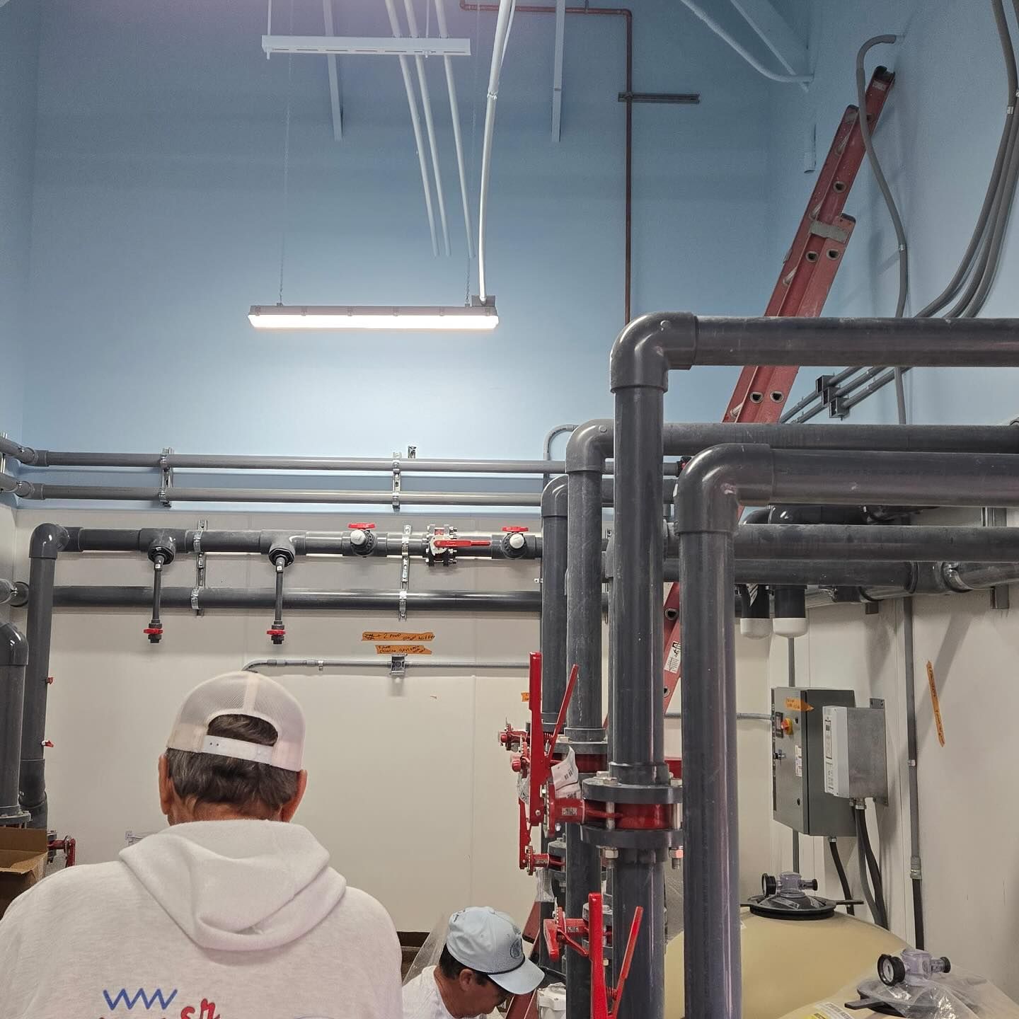 Men in a room with plumbing. One man is wearing a white hat and hoodie, another is working with a pipe.