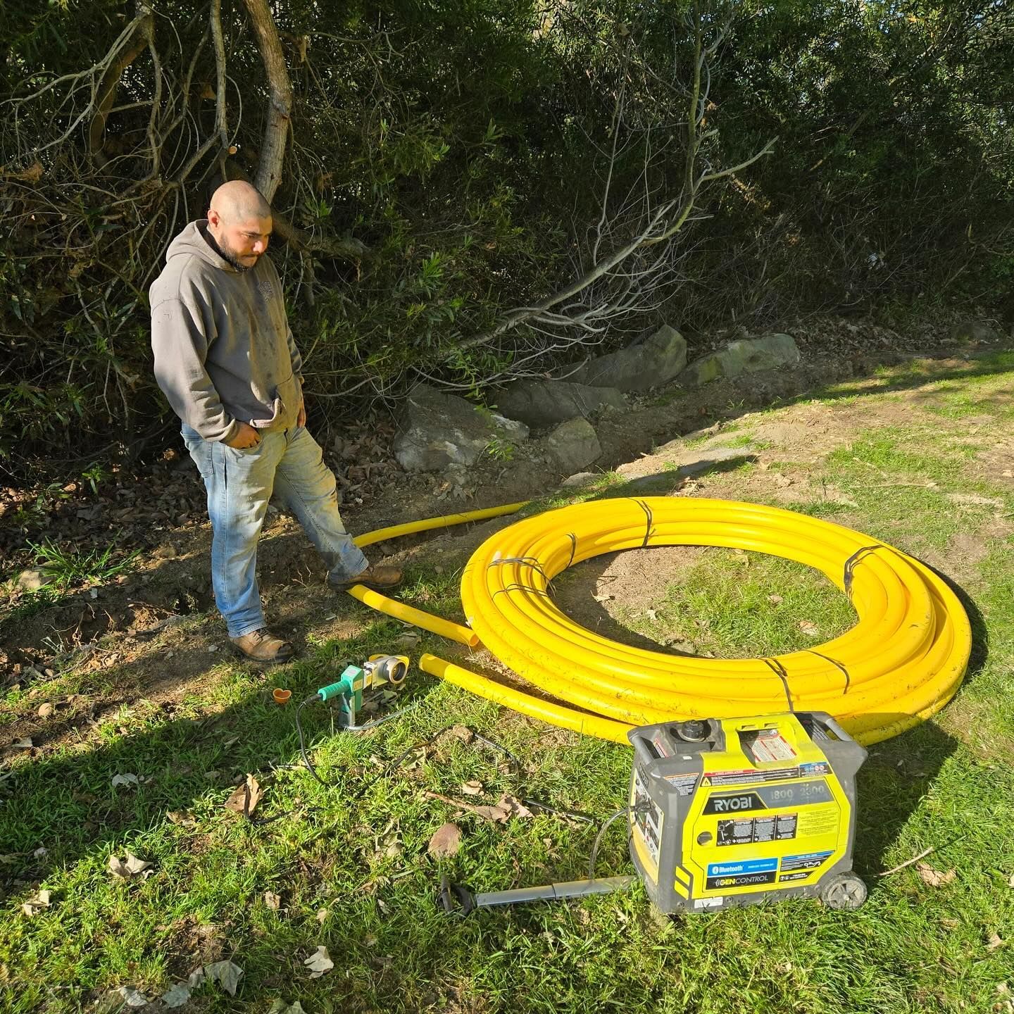 Man standing near coiled yellow pipe, connected to a generator, likely for utility work outdoors.