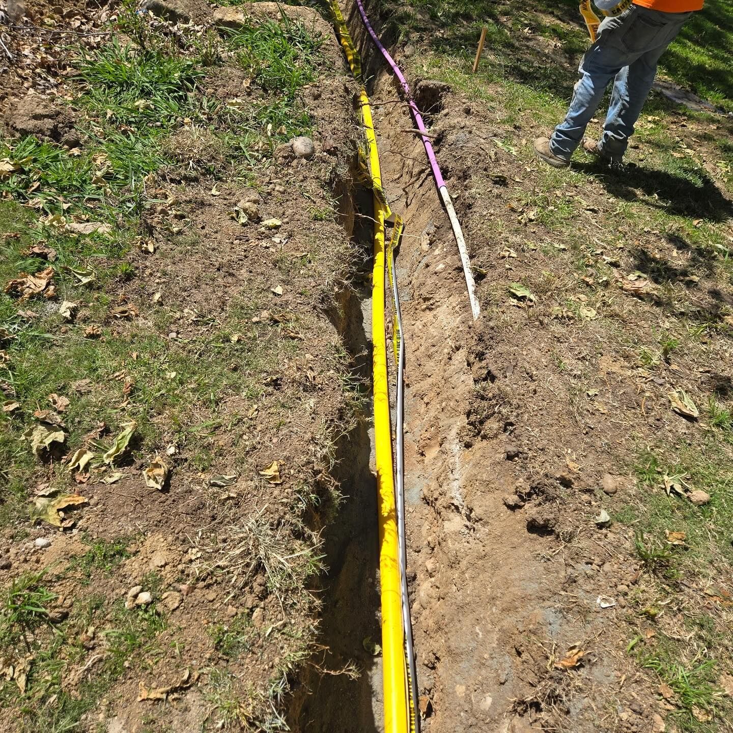 Trench in dirt with yellow pipe and worker's legs.