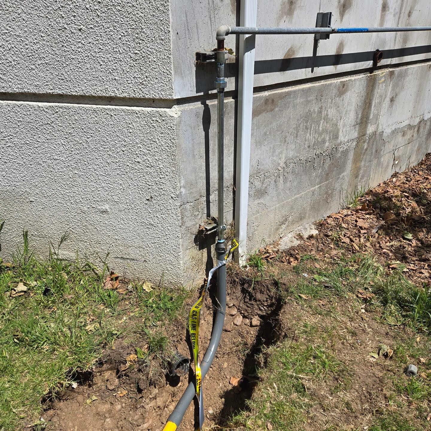 Exterior gas line with exposed black pipe in a trench next to a building wall. Yellow caution tape is visible.