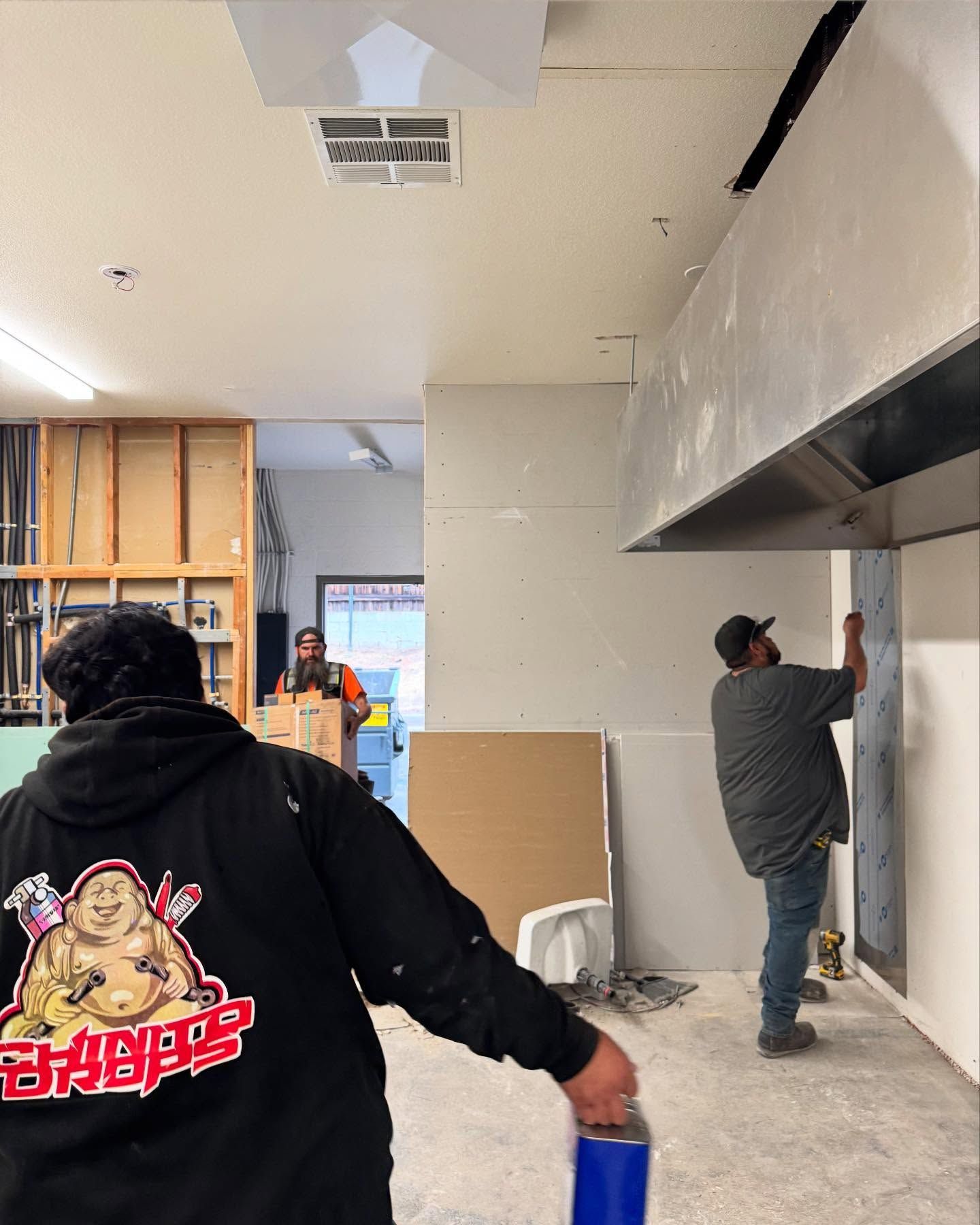 Men installing drywall in a commercial kitchen under construction. One holds a blue container, another works on the wall.