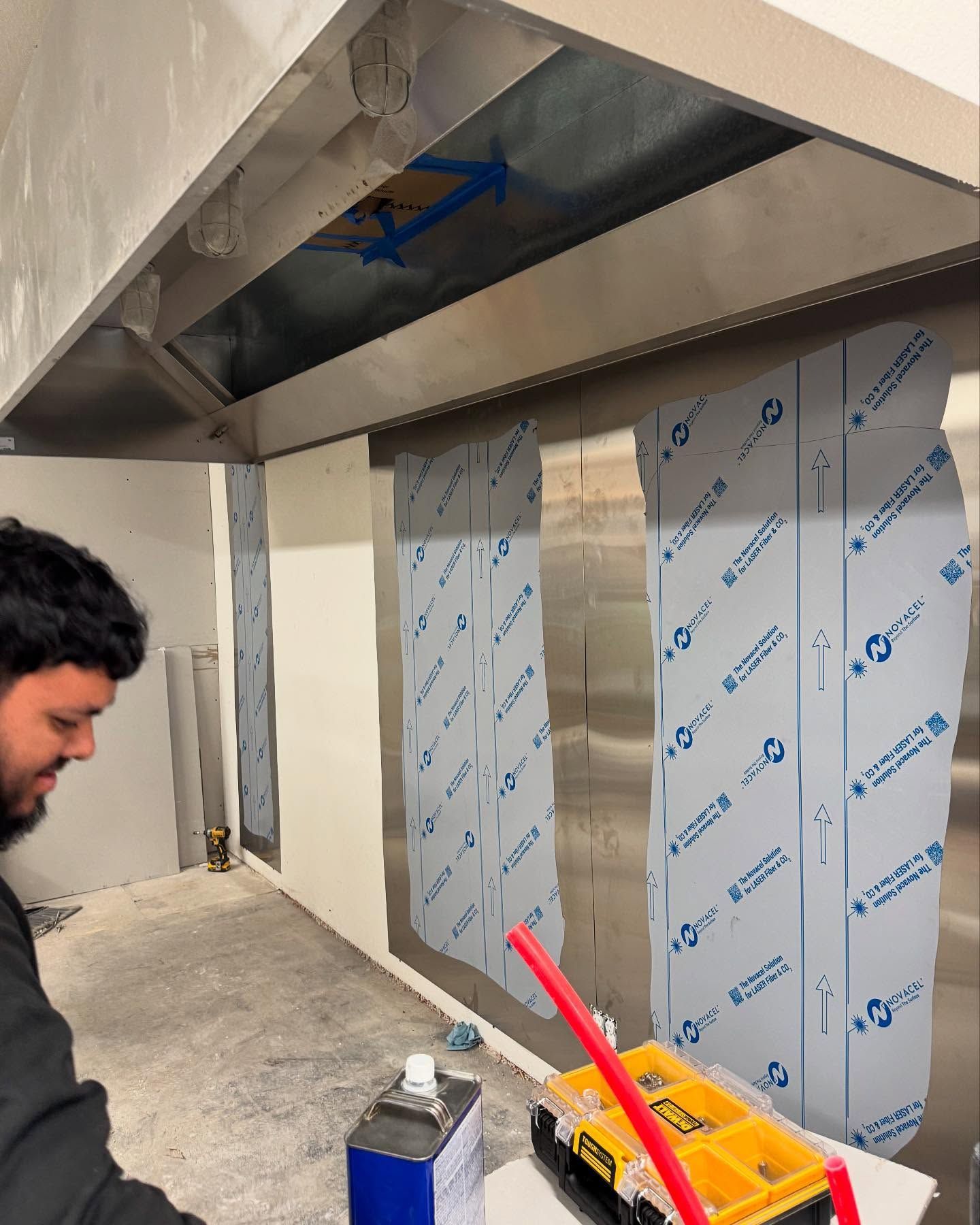 Man installing a stainless steel range hood in a commercial kitchen, with protective film still on the walls.