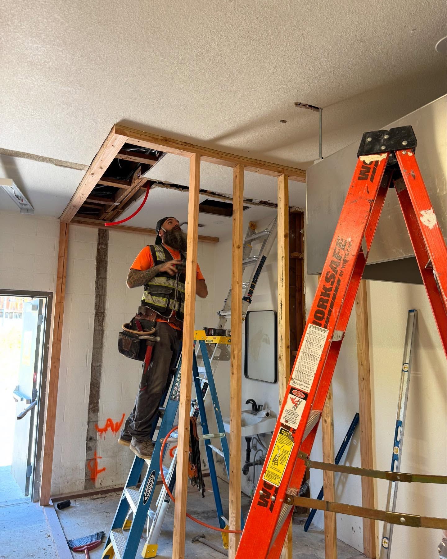 Construction worker framing wall, standing on a ladder. Orange ladder and tool belt visible. Interior setting.
