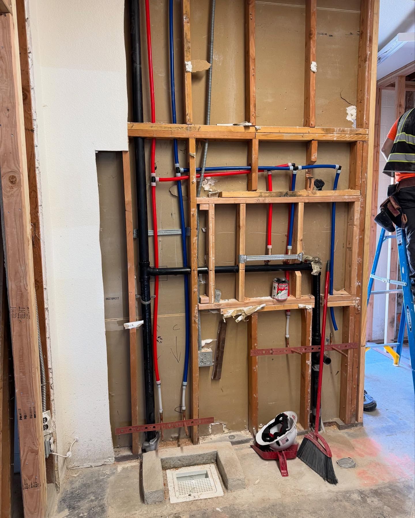 Bathroom wall under construction, plumbing visible. Red, blue pipes, studs, and concrete floor.