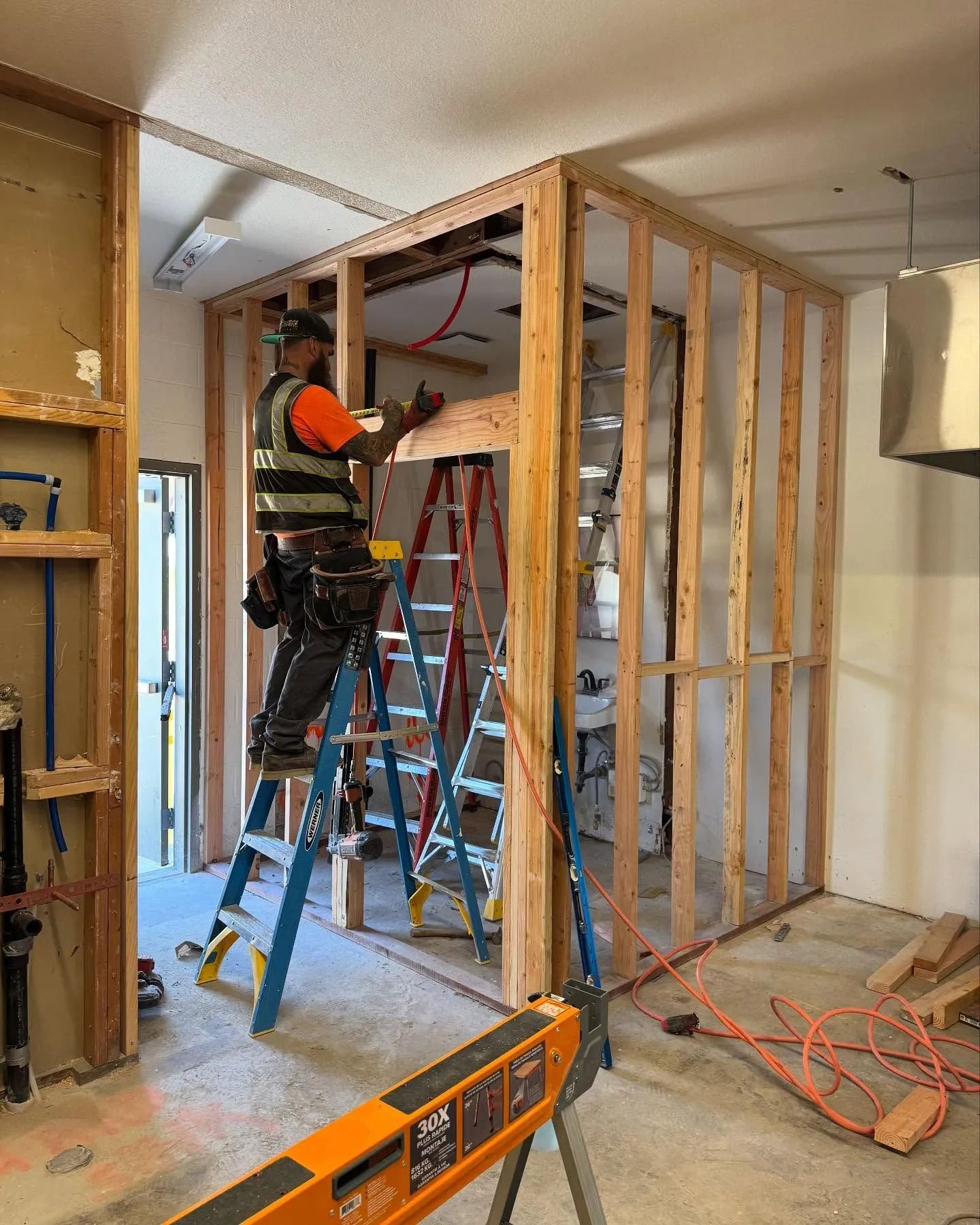 Worker on a ladder framing a wall in an unfinished room with exposed studs and tools.