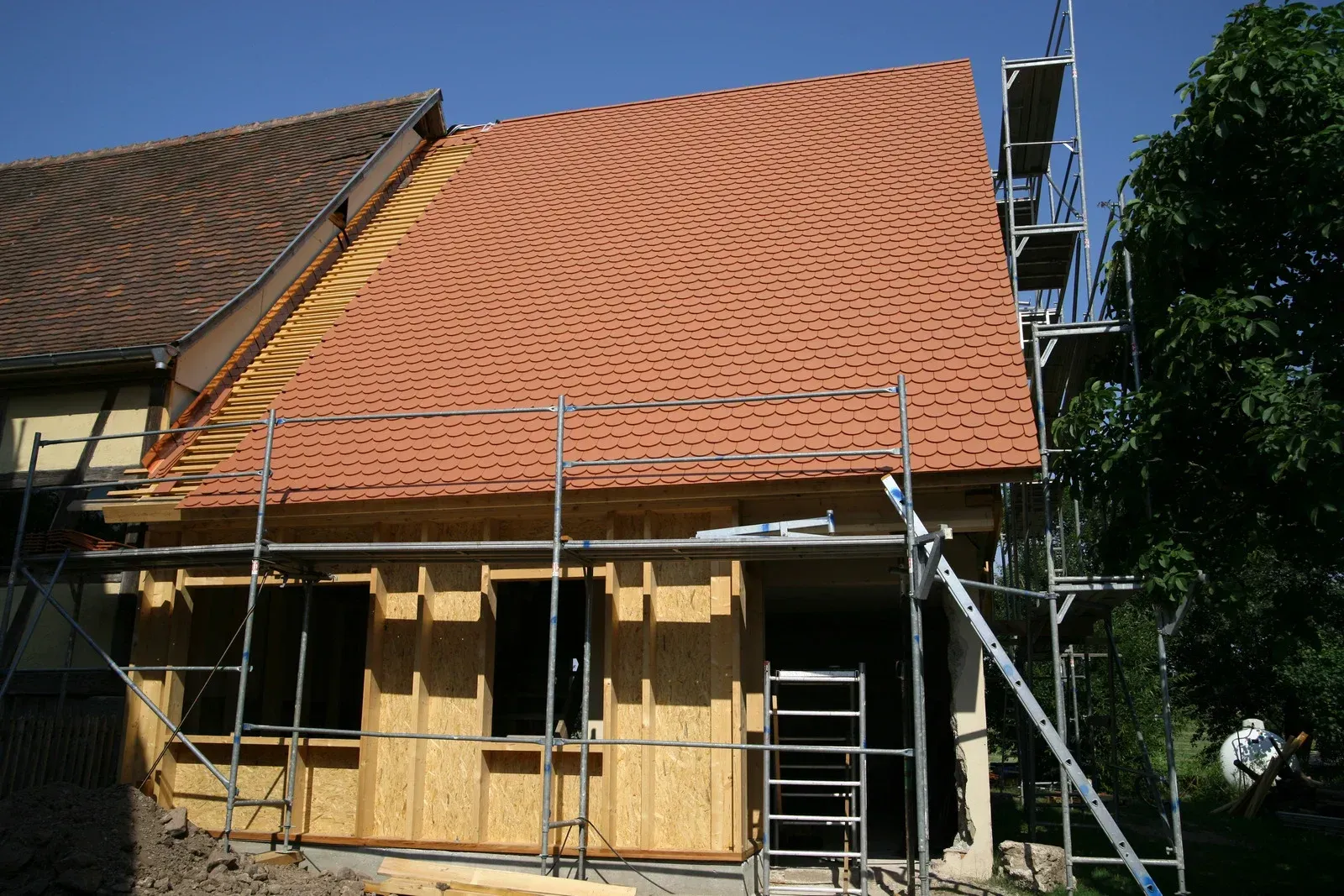Construction of a house addition with a red tile roof, scaffolding, and wooden walls.