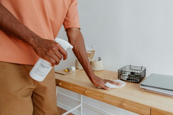 Person cleaning a wooden desk with a spray bottle and cloth in a bright room