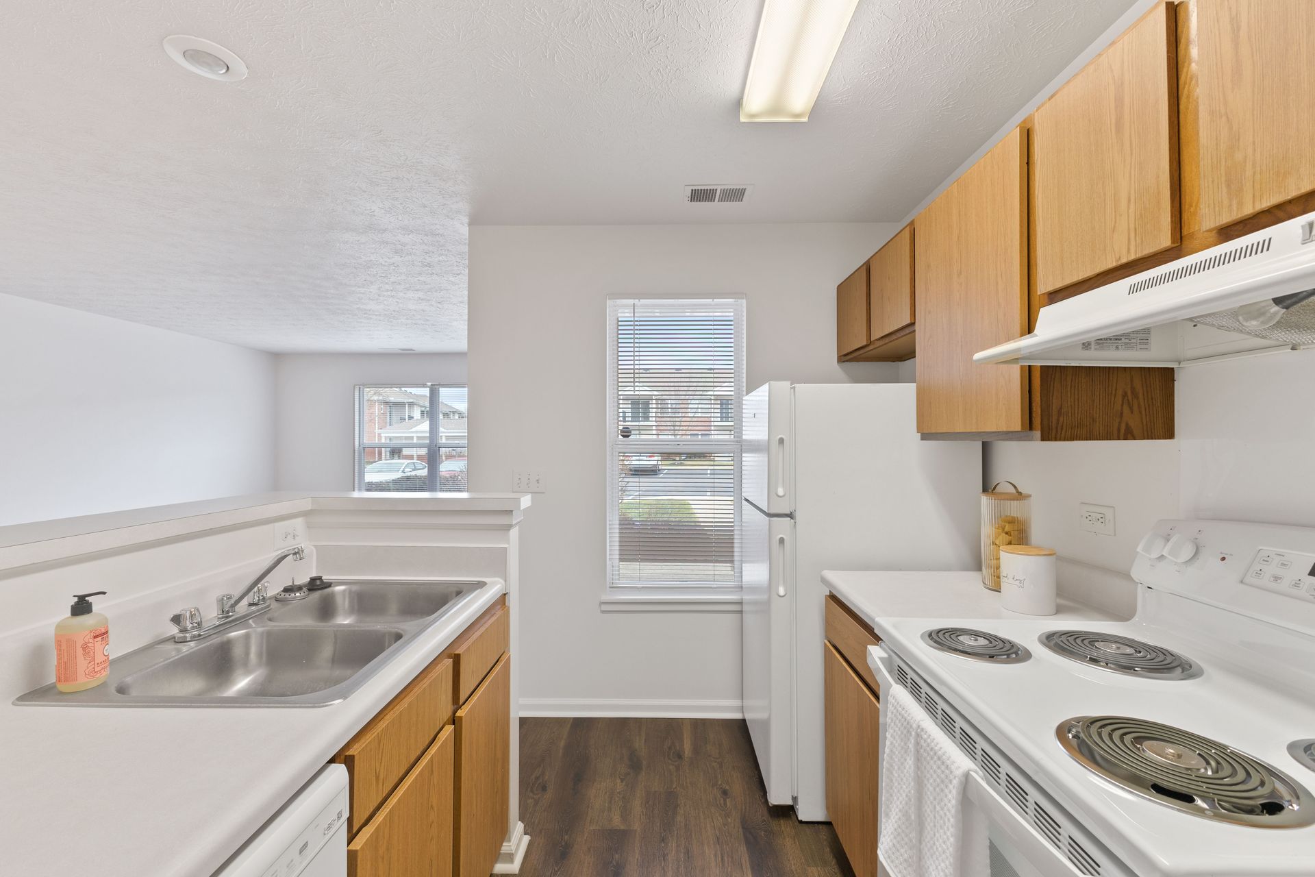 The Maple floor plan kitchen with white appliances, wood cabinetry, and bright window light.