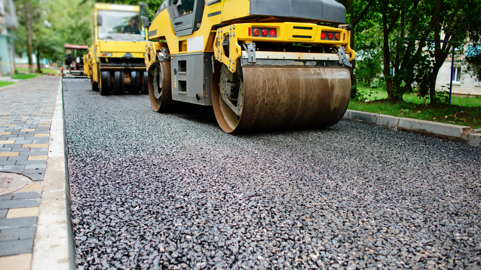 Workers use tools to finish the edges of a newly paved asphalt driveway next to a house on a sunny day.