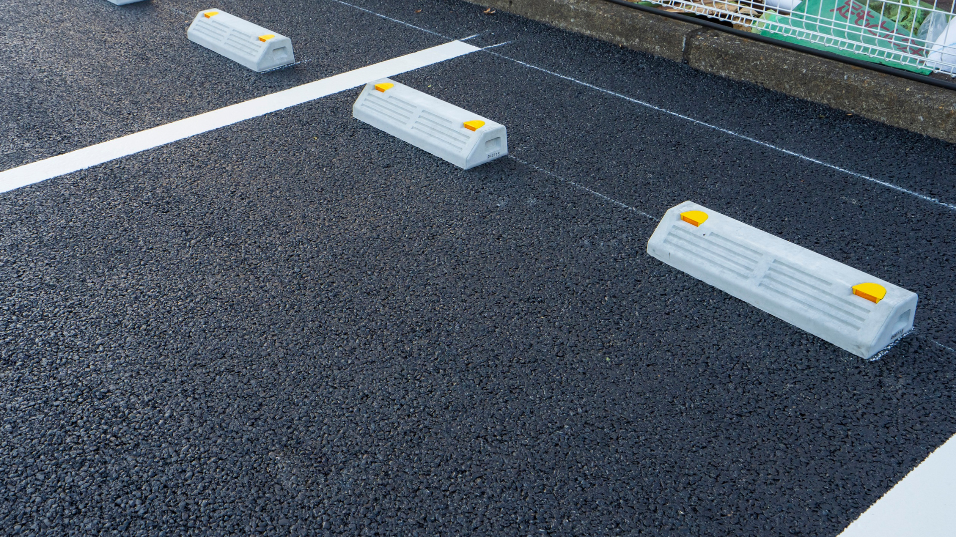 A newly paved asphalt driveway borders a stepped stone retaining wall next to a light-colored house with AC units.