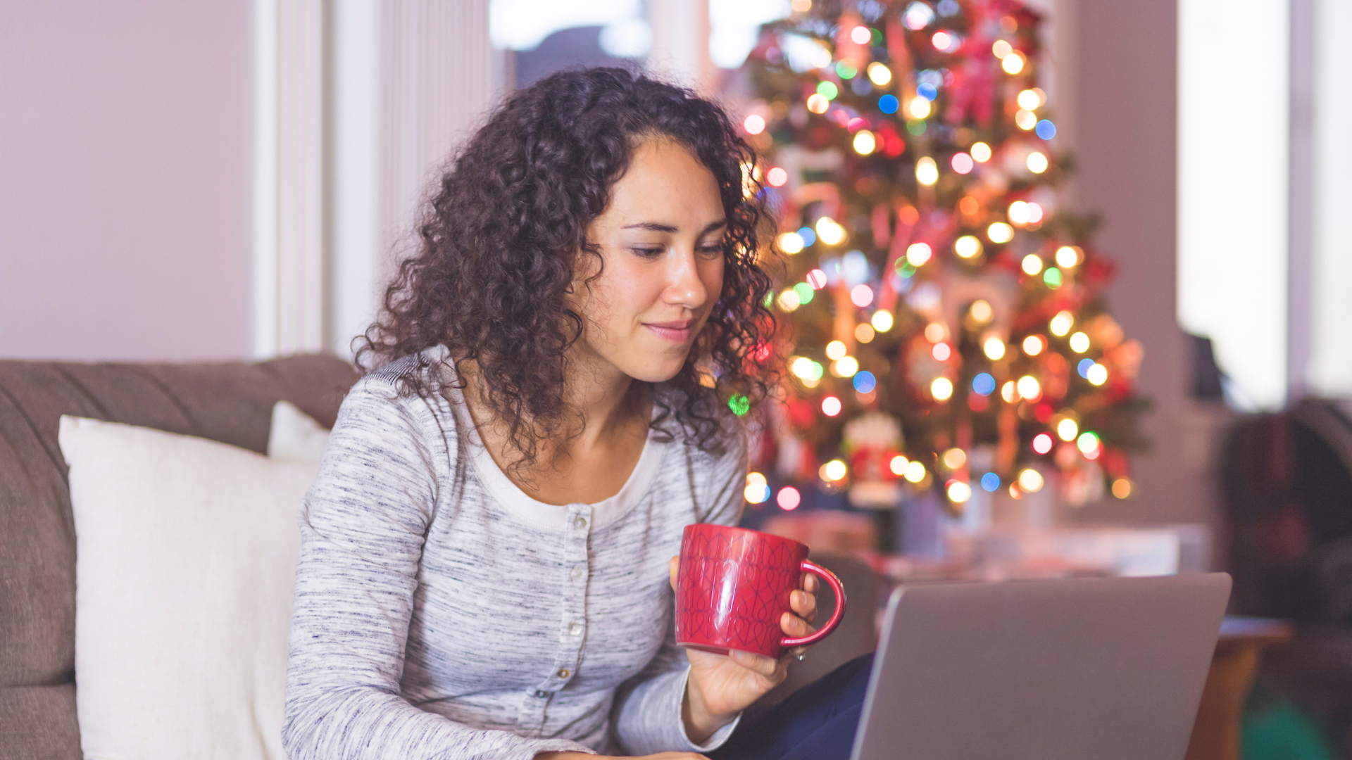 Woman seated on a couch holding a red mug, looking at a laptop with a lit Christmas tree in the background.