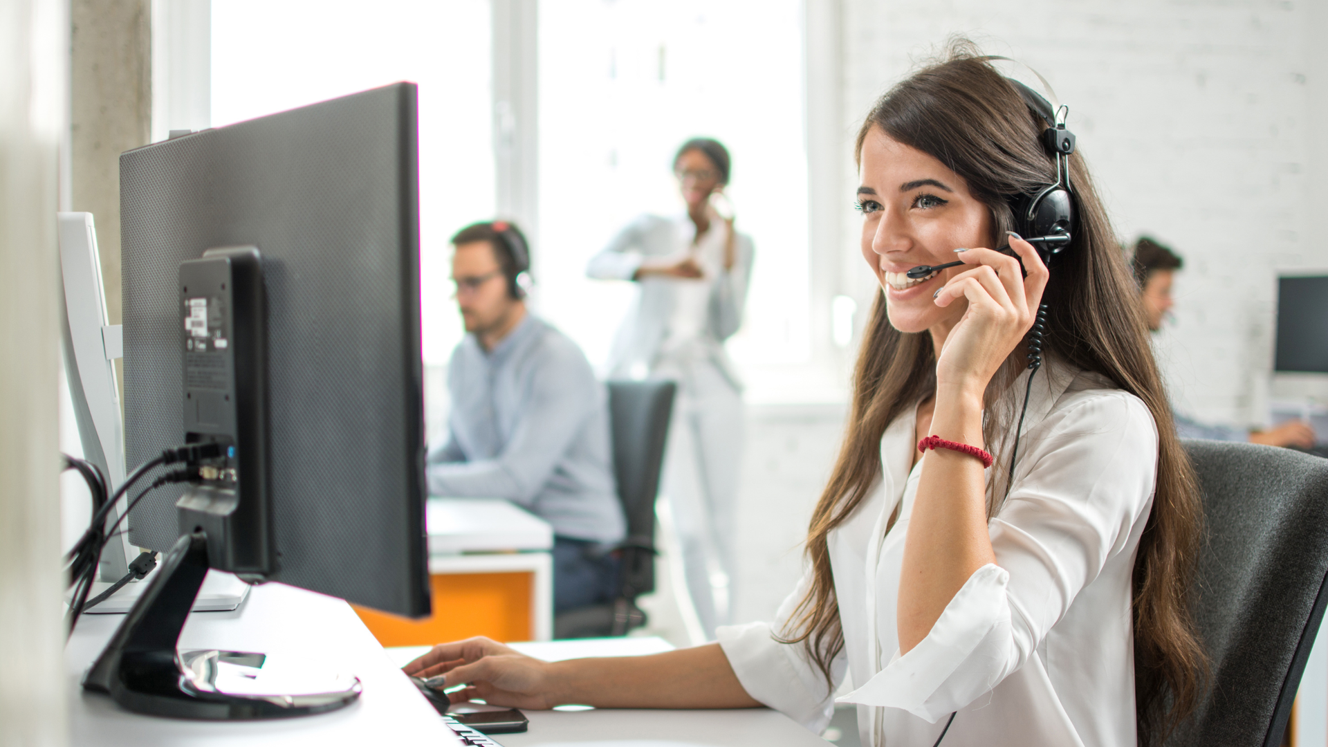 Woman with headset smiles while using a computer in an office setting.