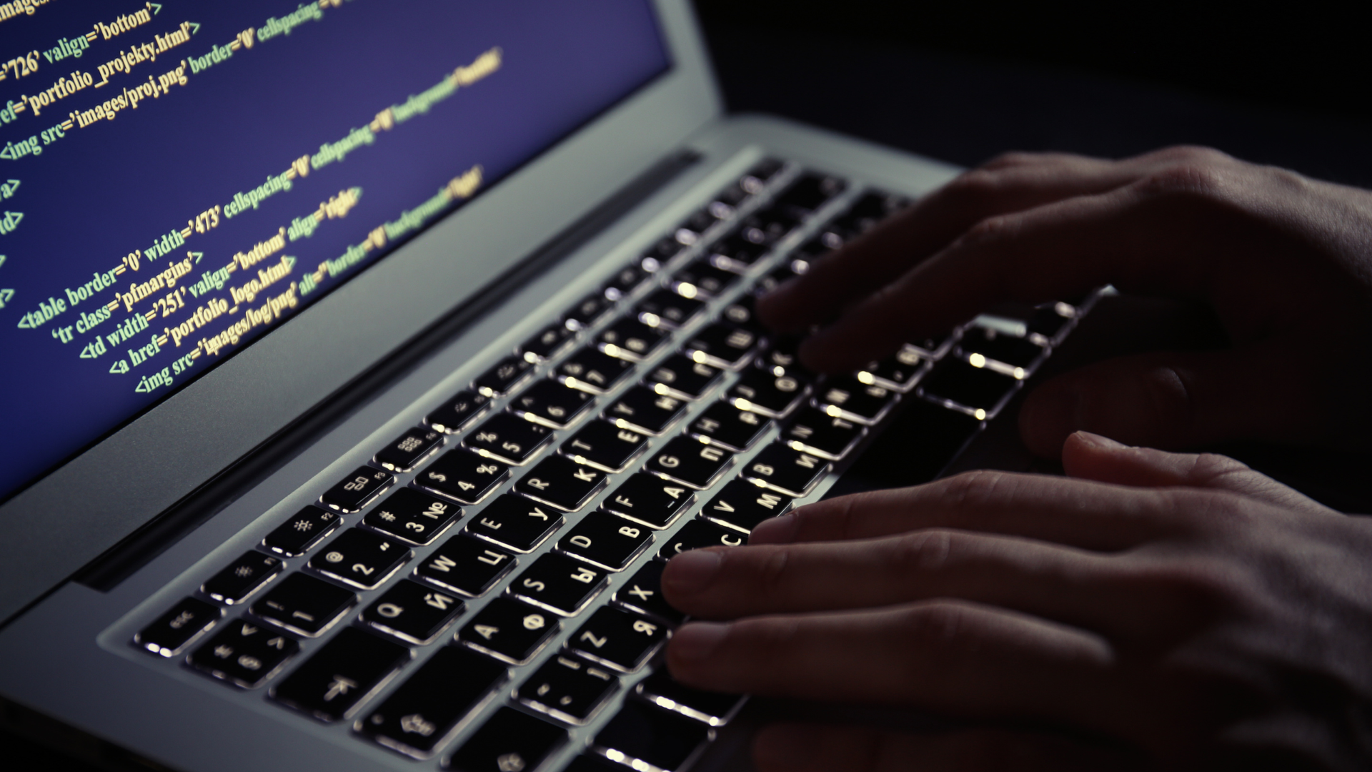 Hands typing on a laptop keyboard, illuminated by the glowing screen displaying lines of code.