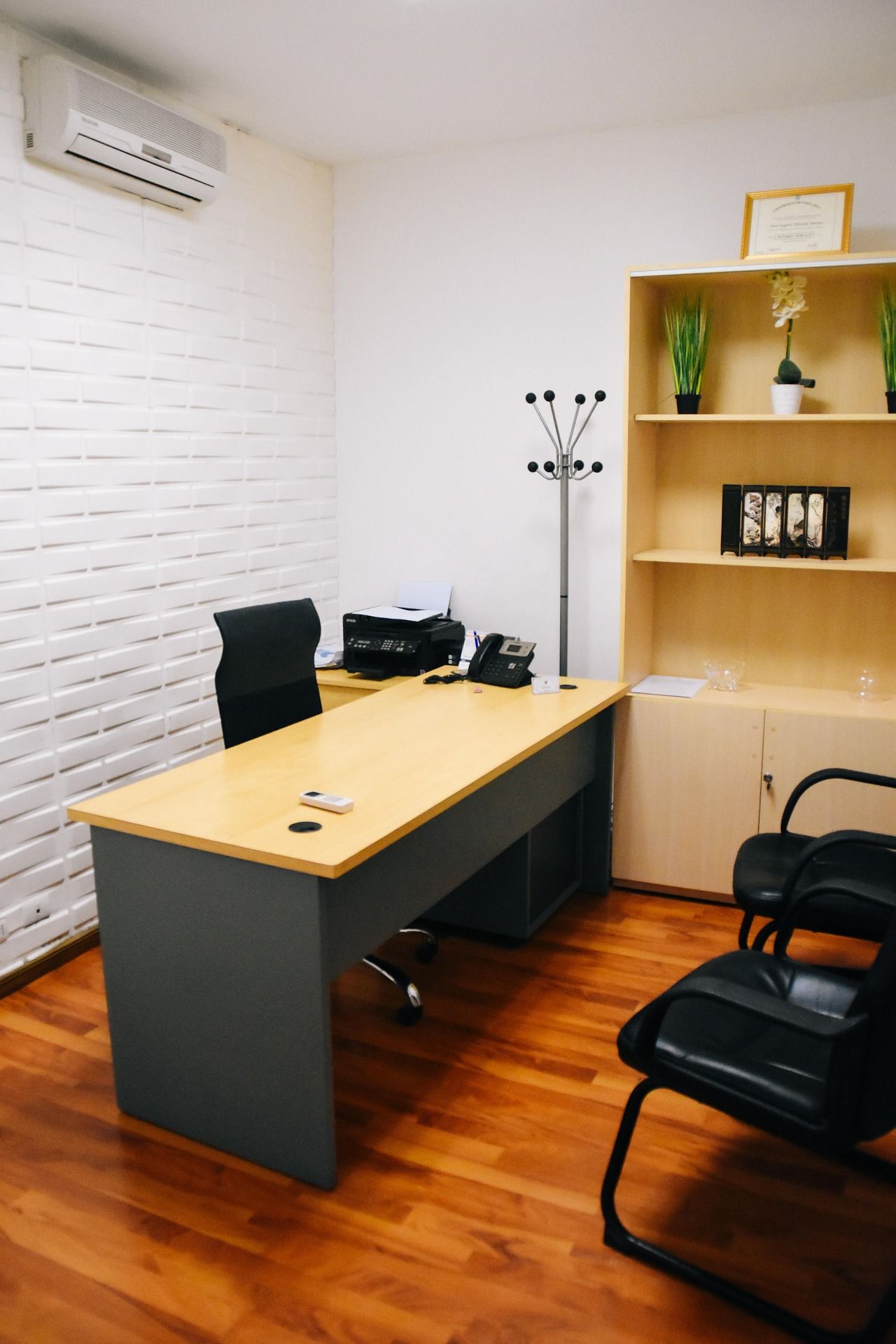 Office with desk, chair, shelving unit, and coat rack. Wooden floor and white brick wall.