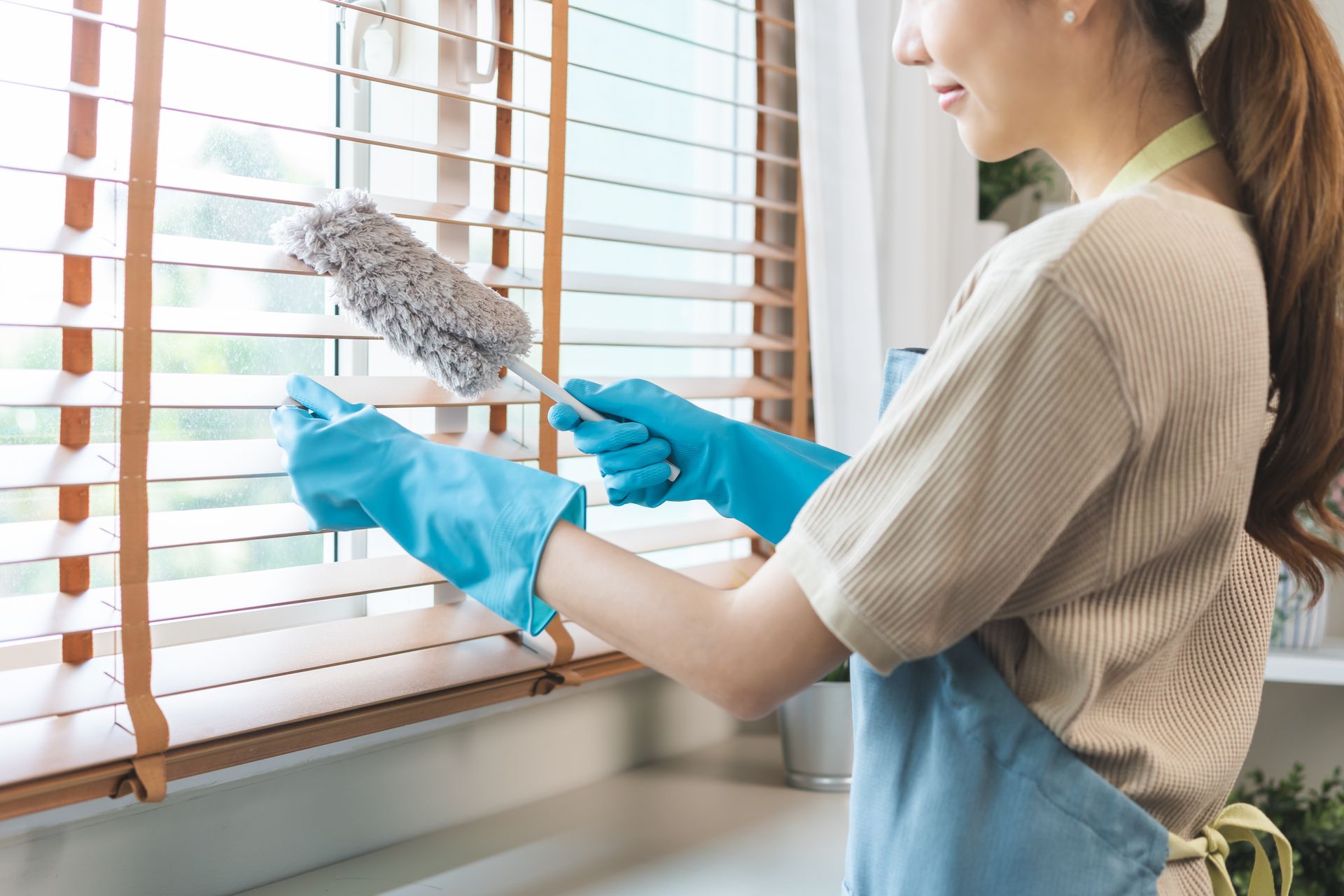 Woman dusting window blinds with a microfiber duster while wearing blue gloves and an apron.