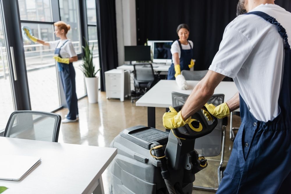 Group of cleaners, wearing aprons and blue gloves, cleaning windows in a bright office.