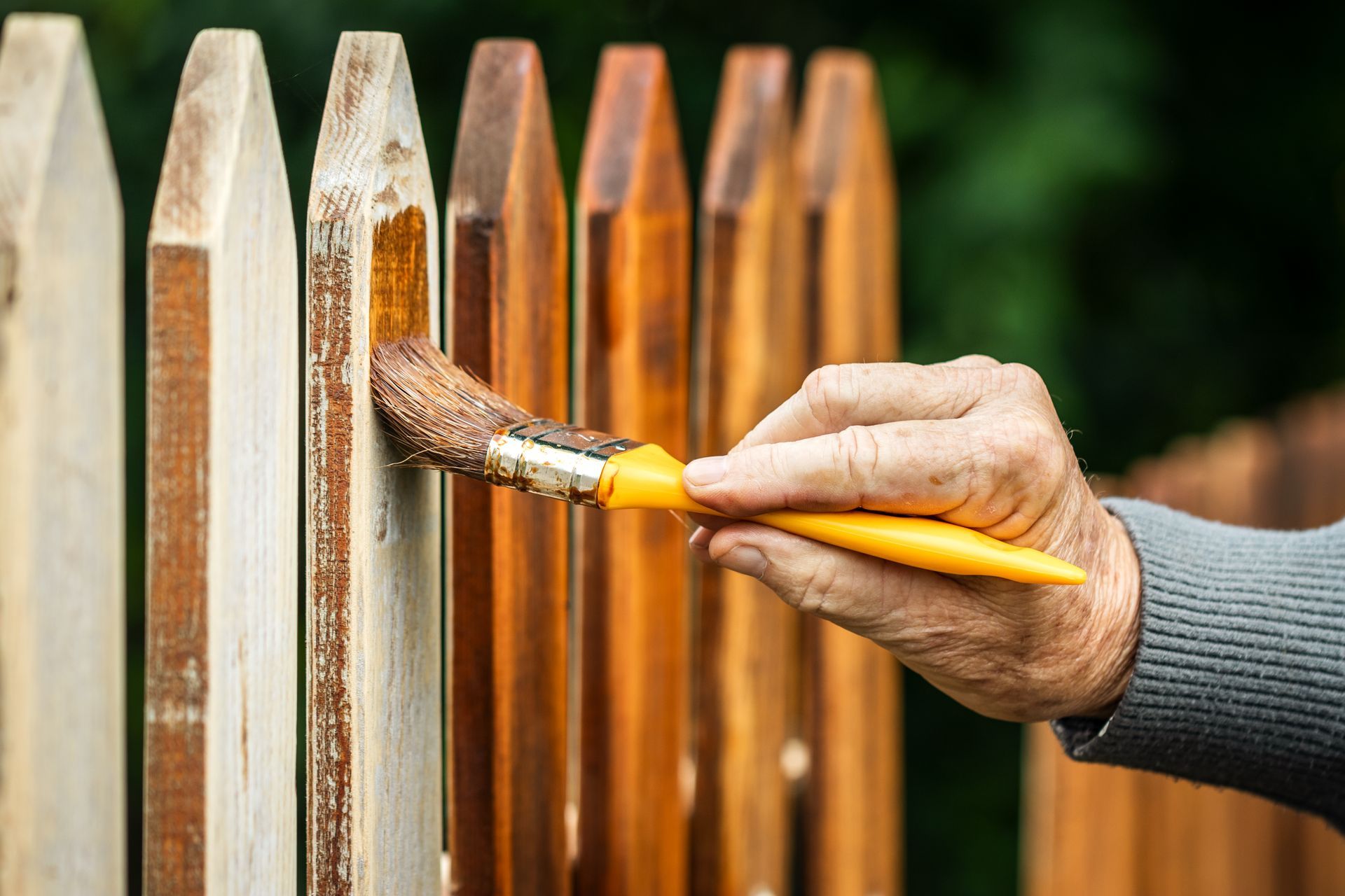 Person painting wooden fence brown with a brush, outdoors.