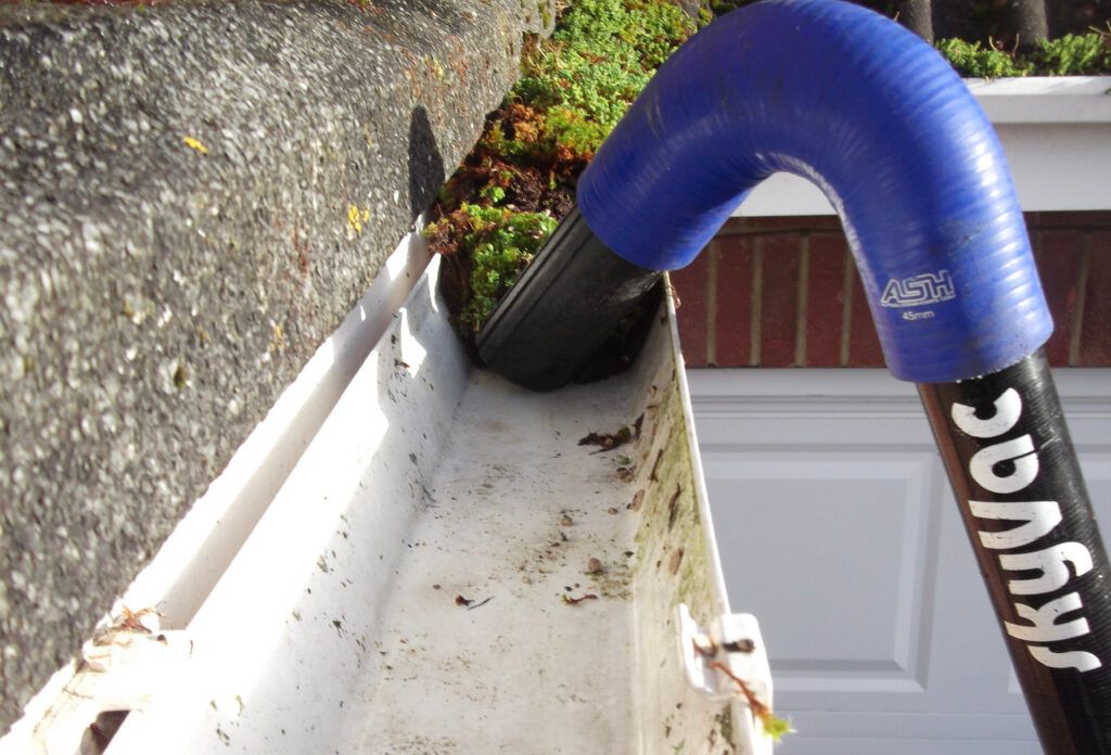 A person using a SkyVac to clean a gutter. The vacuum has a blue flexible hose and a black nozzle.