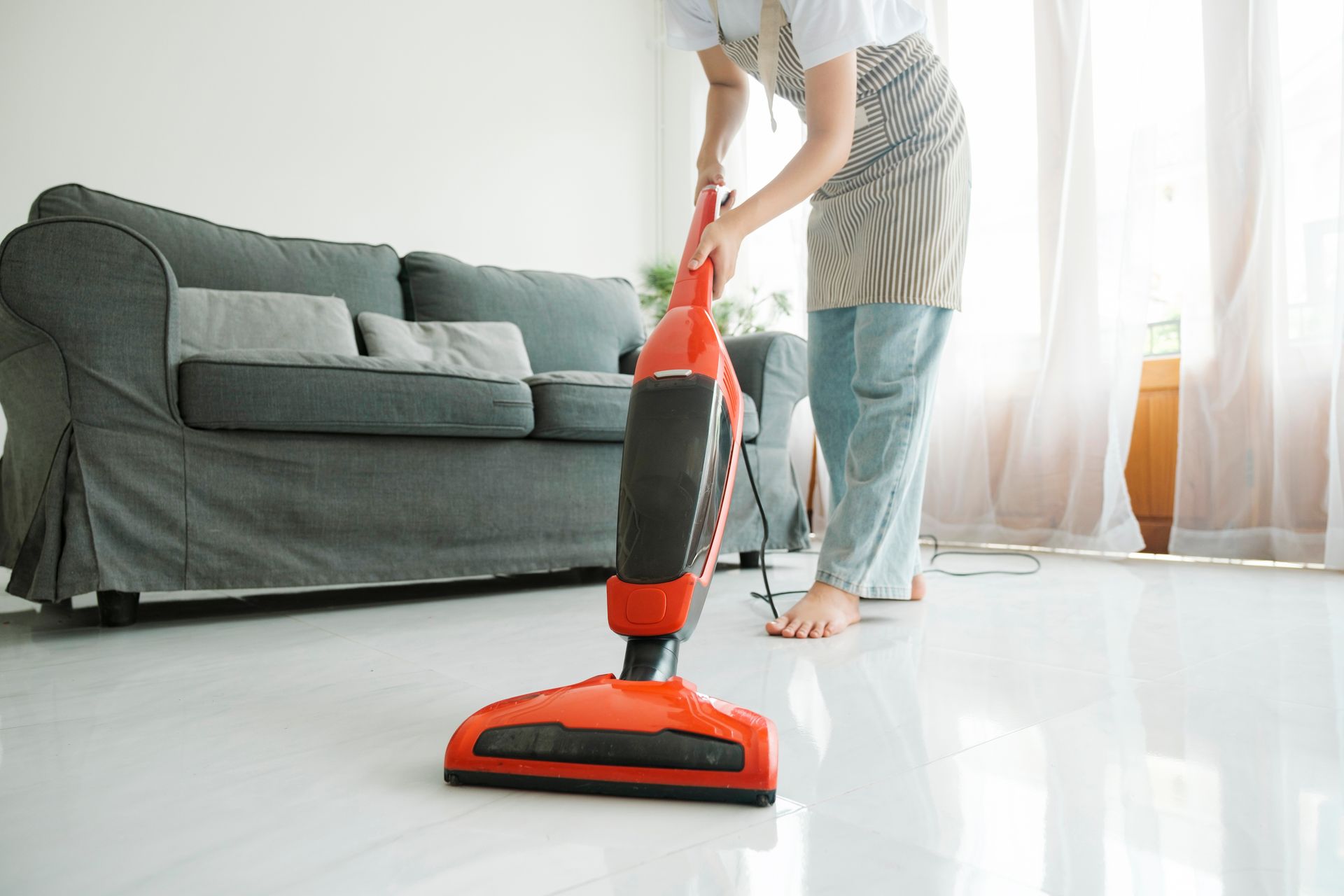 Woman vacuuming a shiny, white floor in a living room with a gray couch and sheer curtains.