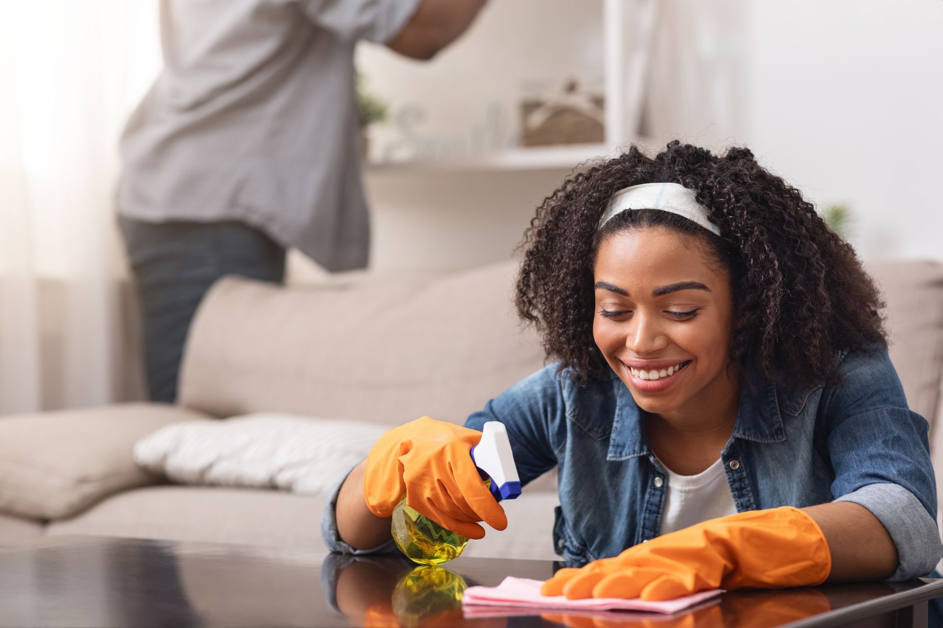 Woman wearing orange gloves cleaning a table with a spray bottle and cloth; another person in background.