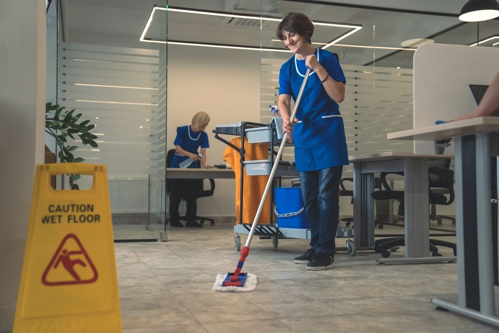 Group of cleaners, wearing aprons and blue gloves, cleaning windows in a bright office.