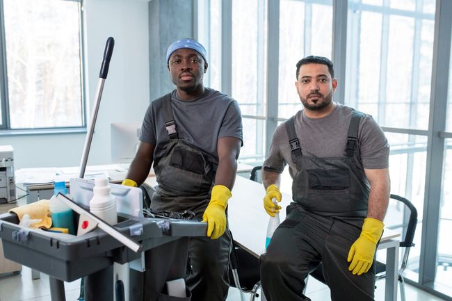 Two men in work clothes and gloves stand near a cleaning cart in an office.