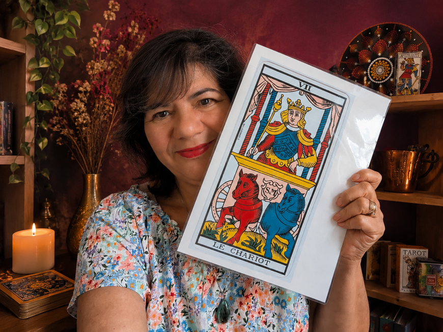 Une femme portant des lunettes tient deux cartes de tarot sur une table où se trouvent également d'autres cartes de tarot, des bougies et une pancarte indiquant qu'il s'agit d'une séance de tirage de tarot.