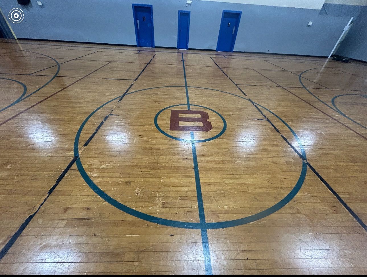 Gymnasium hardwood floor with basketball court markings; three blue doors in the background.