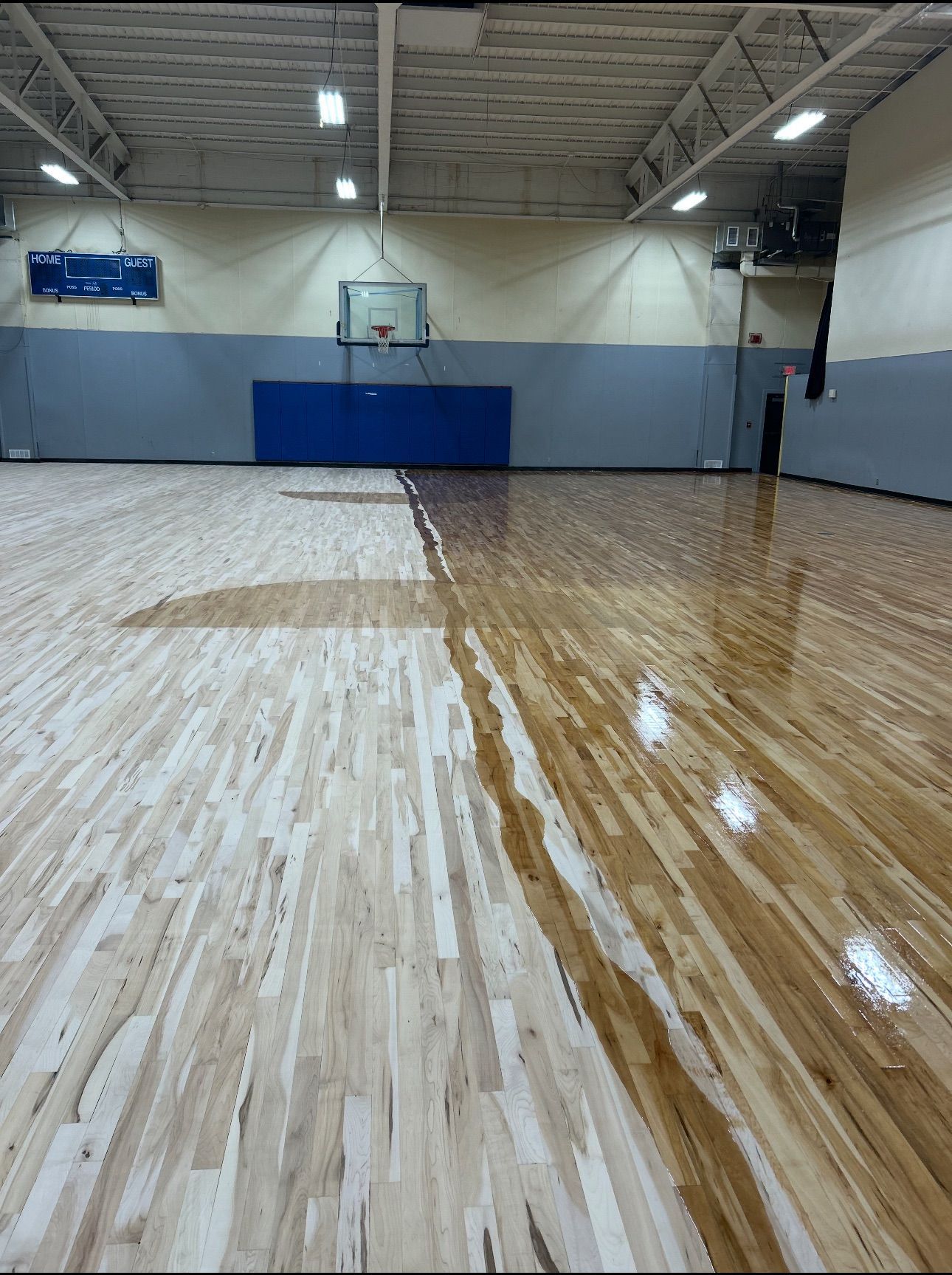 Basketball court with wet, varnished wood floor. Blue wall pads, hoop, and scoreboard in the background.
