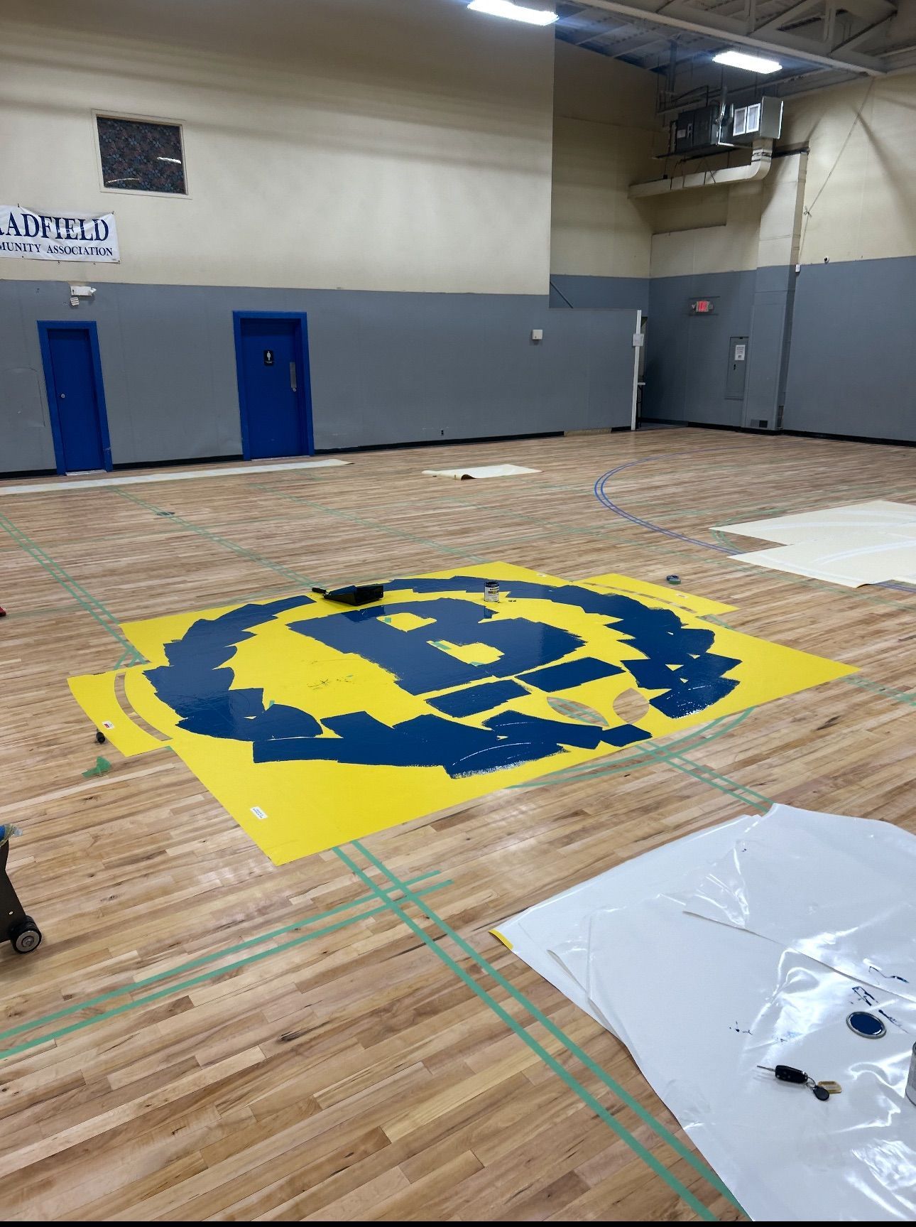 Basketball court with large yellow and blue emblem being renovated. Blue doors and gray walls in background.