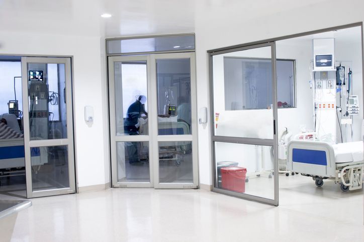 Hospital corridor with glass doors, patient beds visible inside rooms, a medical setting.