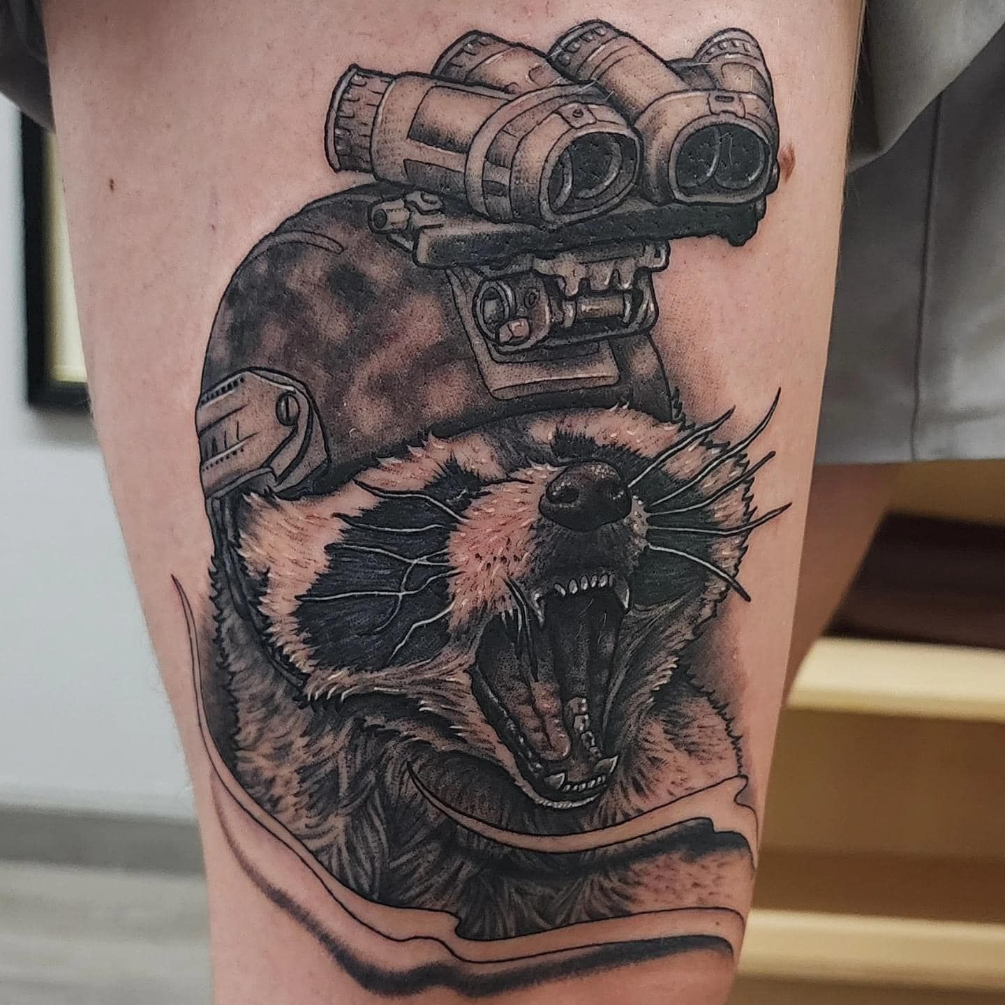 Black-and-gray tattoo of a roaring raccoon wearing night-vision goggles on a forearm
