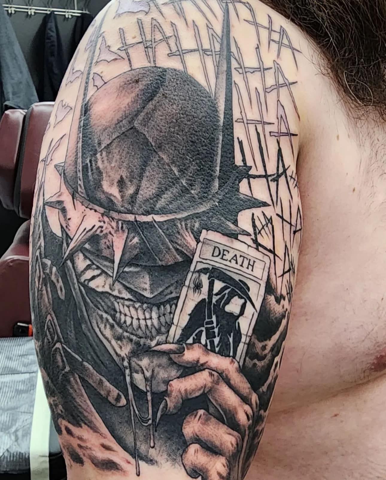 Close-up of a black-and-gray skull face tattoo with a cracked visor and a hand holding a “Death” card.