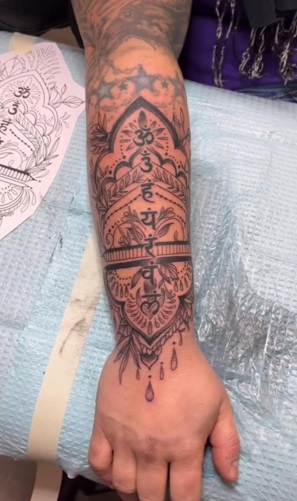Tattooed forearm resting on a medical pad, showing intricate black linework and a dangling red bead design