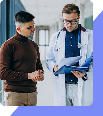 A doctor is talking to a patient in a hospital hallway while looking at a clipboard.