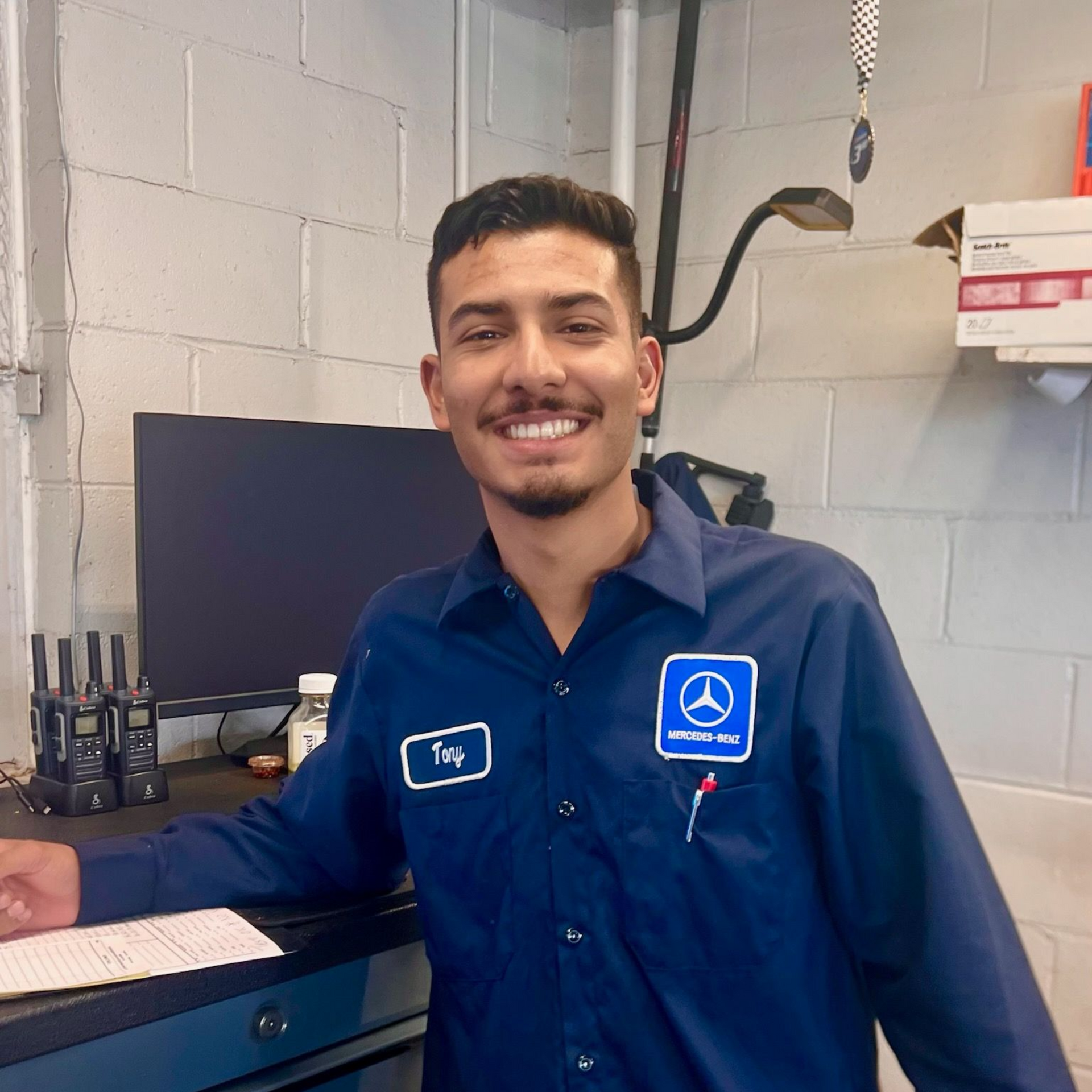 Smiling mechanic in blue uniform at a Mercedes-Benz shop, leaning on a desk.