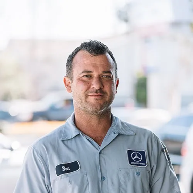 Mechanic in light blue uniform, Mercedes logo on shirt, standing outdoors. Name tag reads