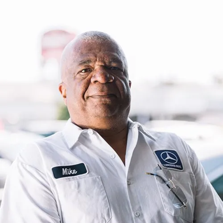 Man in a white uniform with a Mercedes-Benz logo, smiling slightly, outdoors.